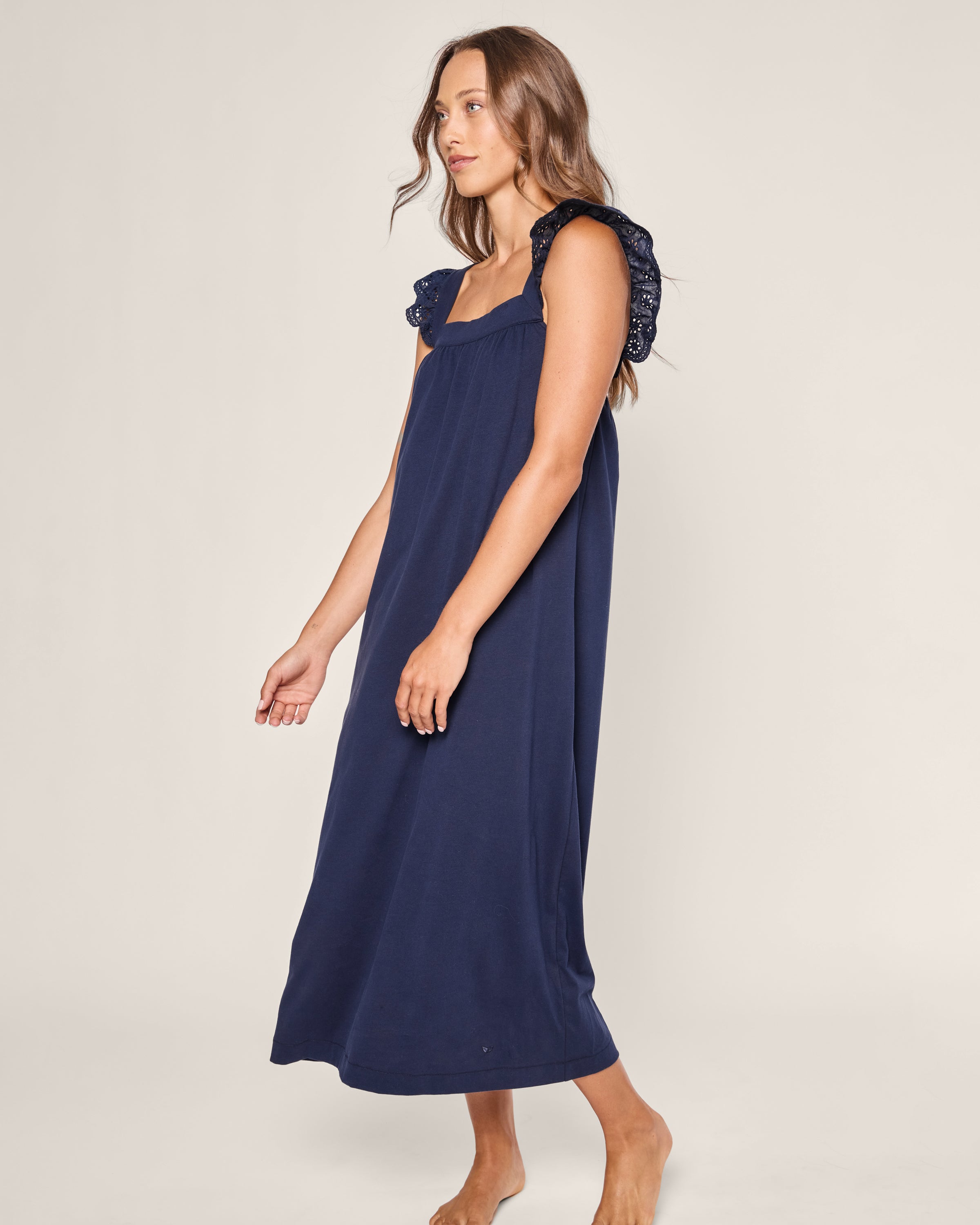 A woman stands barefoot in a Petite Plume Womens Pima Clara Nightgown in navy, her wavy brown hair cascading as she gazes to the side. The light-colored background highlights the elegance of her dress crafted from luxurious Peruvian Pima cotton.