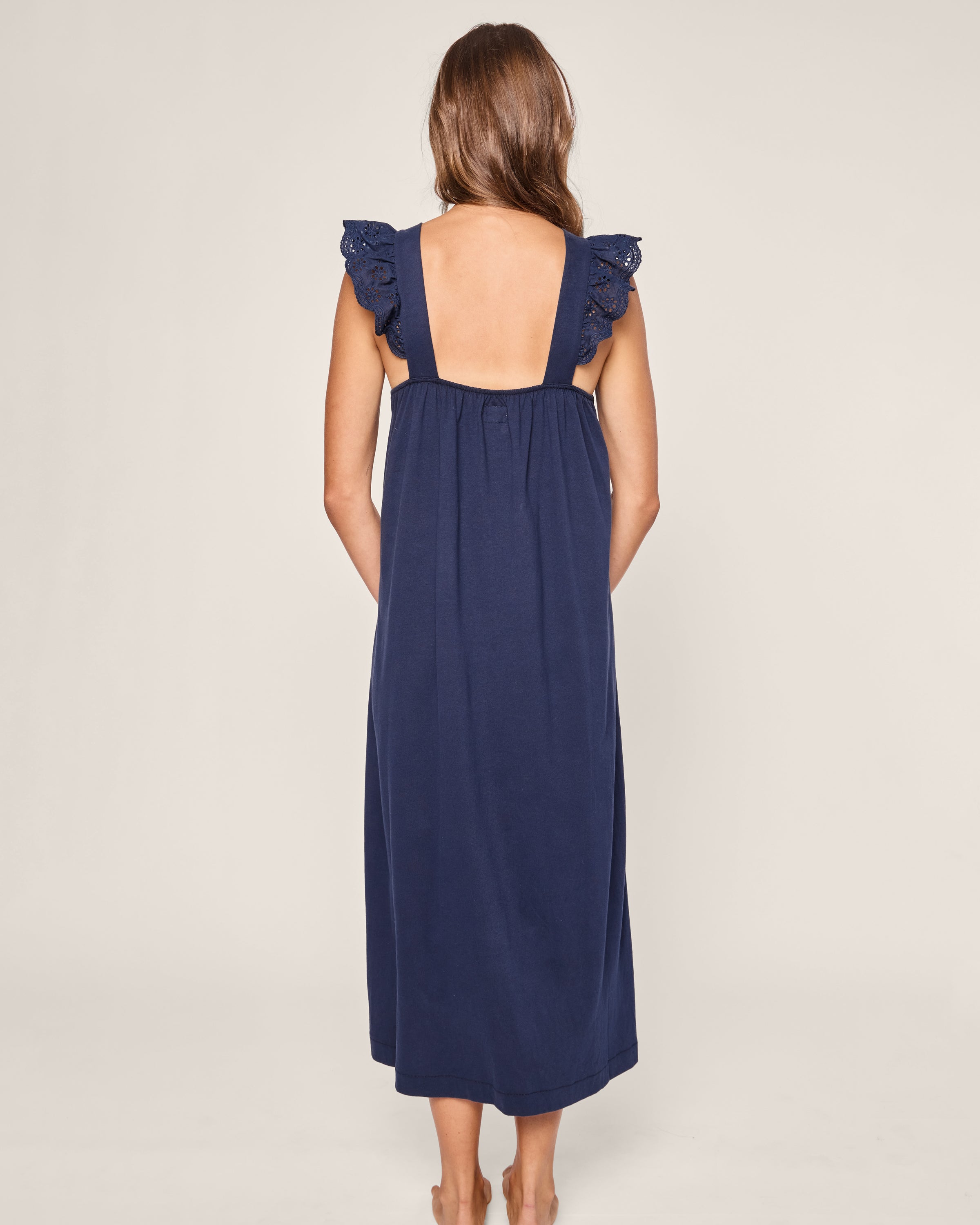 The back view showcases a woman in Petite Plumes Womens Pima Clara Nightgown in Navy, featuring ruffled straps and a luxurious sleepwear design. She stands barefoot against a light background, her long wavy hair gracefully cascading down her back.