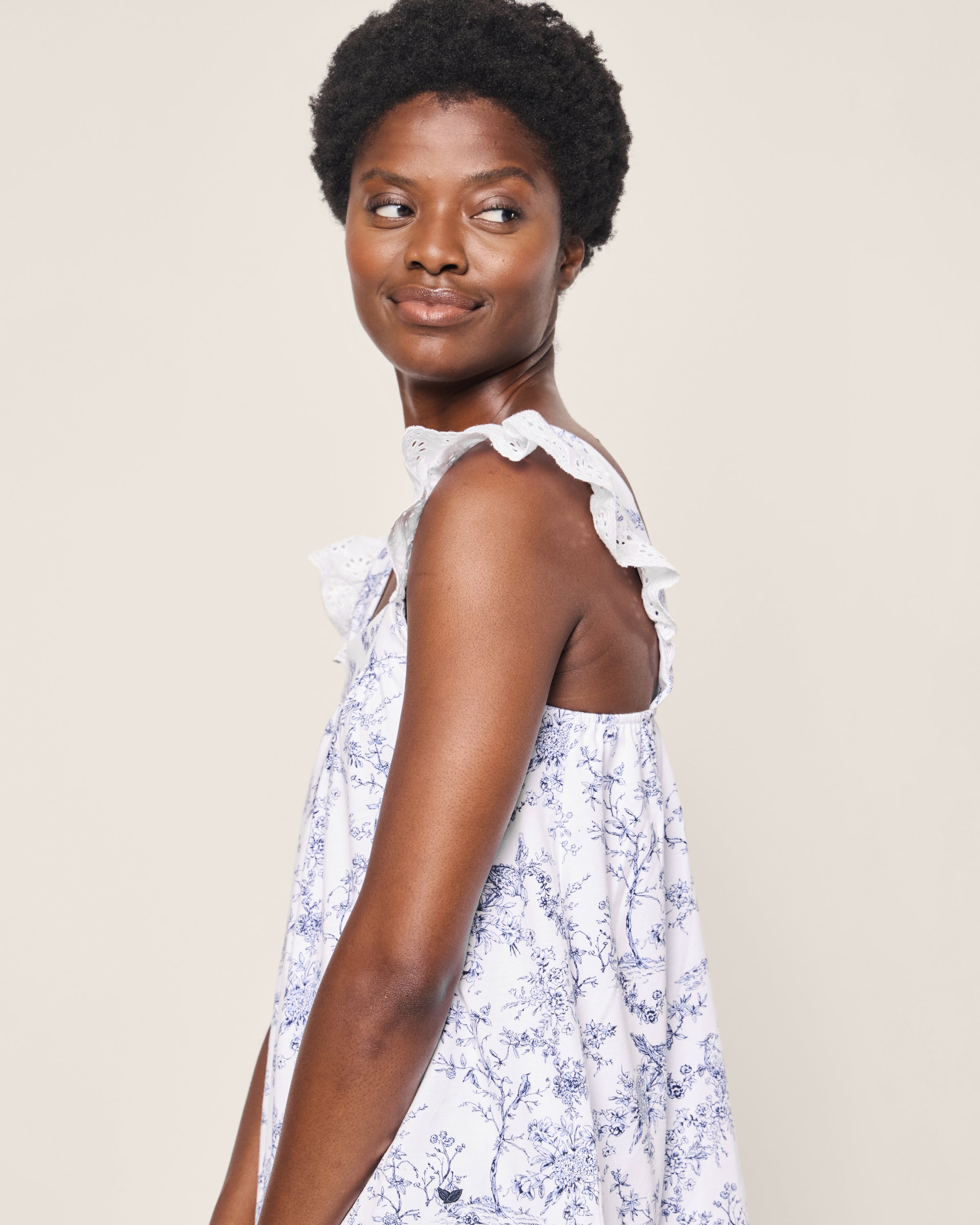 A woman in the Womens Pima Clara Short Set by Petite Plume, featuring a white and blue floral design with ruffled shoulders, exudes botanical elegance as she gazes over her shoulder against a plain background.