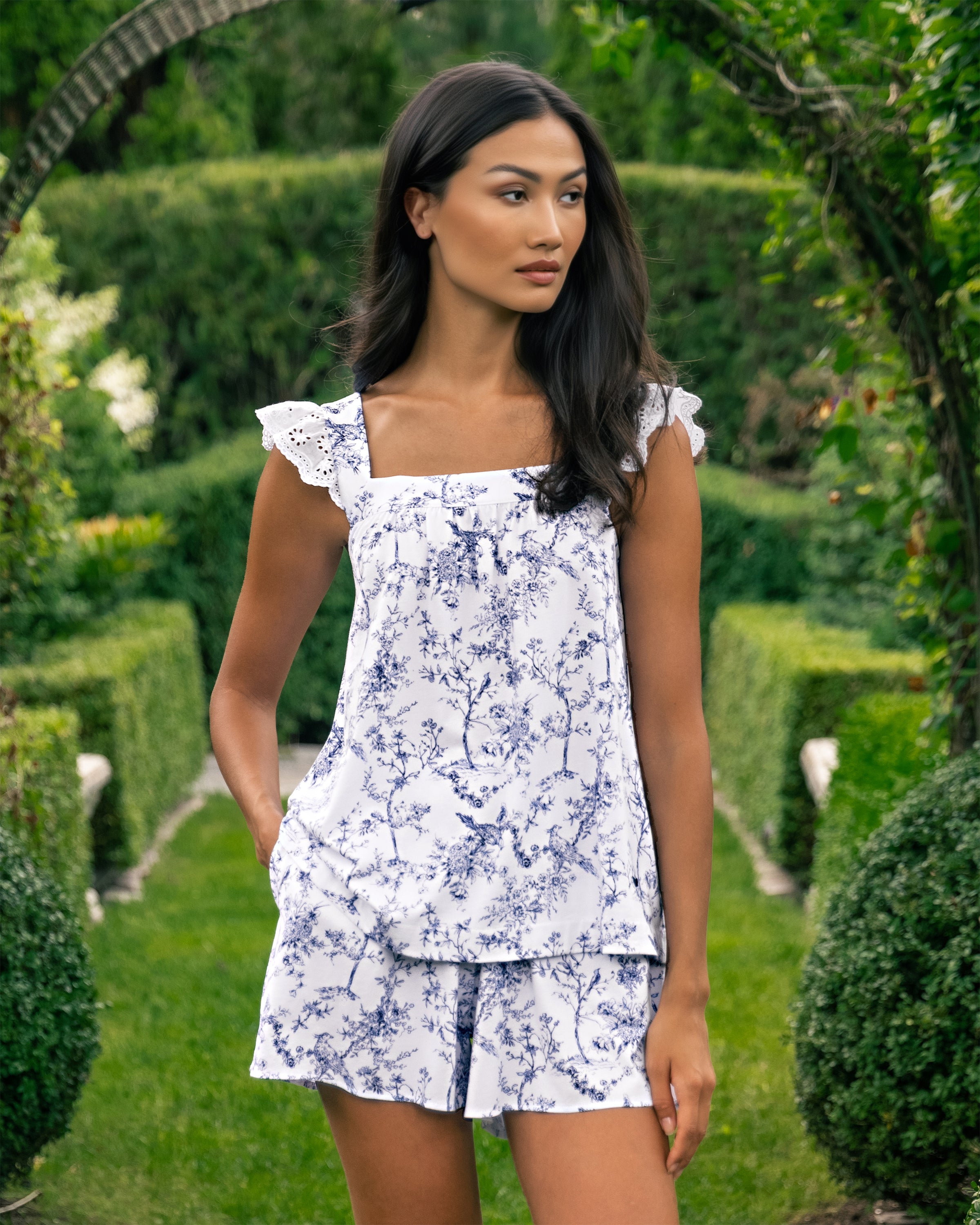 In a garden, surrounded by manicured hedges and an archway, a woman donning the Womens Pima Clara Short Set in Timeless Toile from Petite Plume stands with her hands in her pockets, gazing to the side.