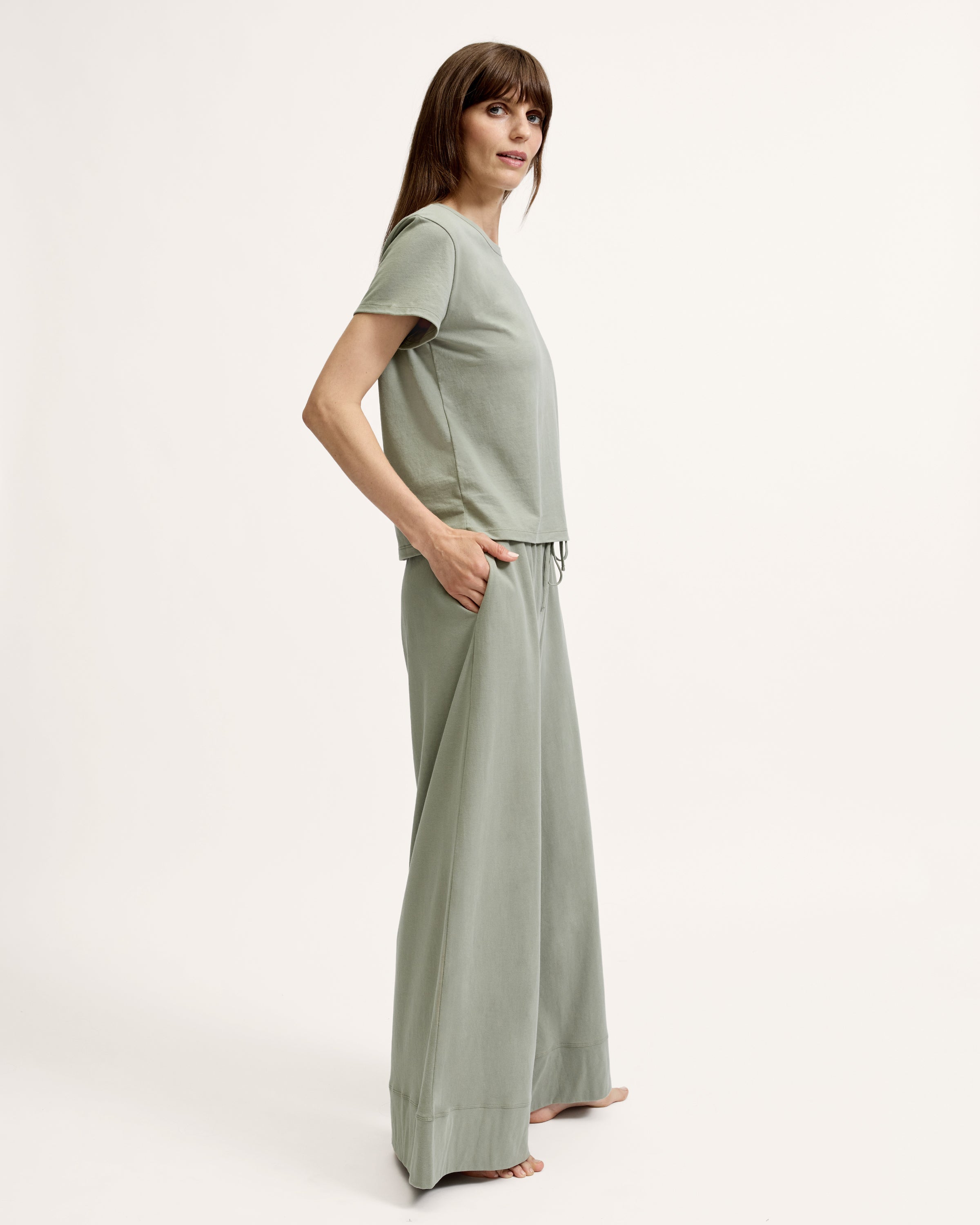 A woman with straight brown hair wears the Women's Pima Essential Crew Neck Tee in Sage and matching drawstring shorts, standing with her hands on her hips against a plain light background.
