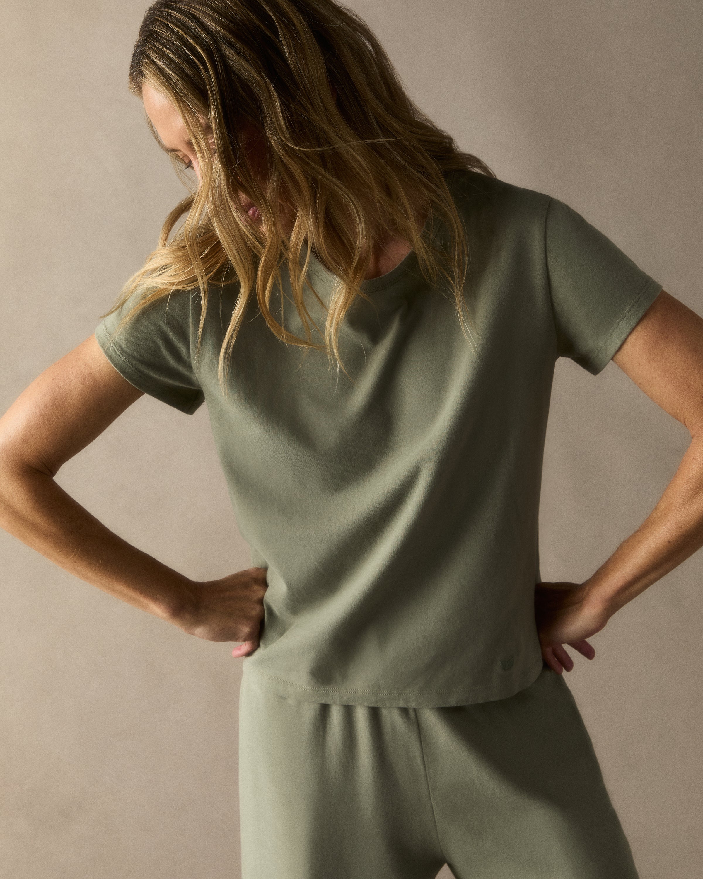 A woman with straight brown hair wears the Women's Pima Essential Crew Neck Tee in Sage and matching drawstring shorts, standing with her hands on her hips against a plain light background.