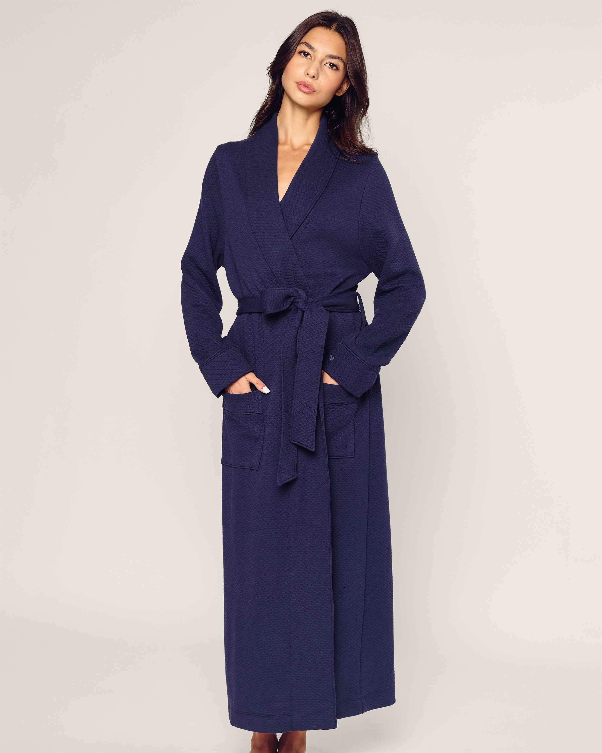 A person in Petite Plumes Womens Pima Ophelia Robe in Blue Dusk Jacquard stands against a plain, light background. The Peruvian Pima cotton robe features a belt and two front pockets, with their hands resting casually inside. They gaze ahead, exuding relaxed sophistication.