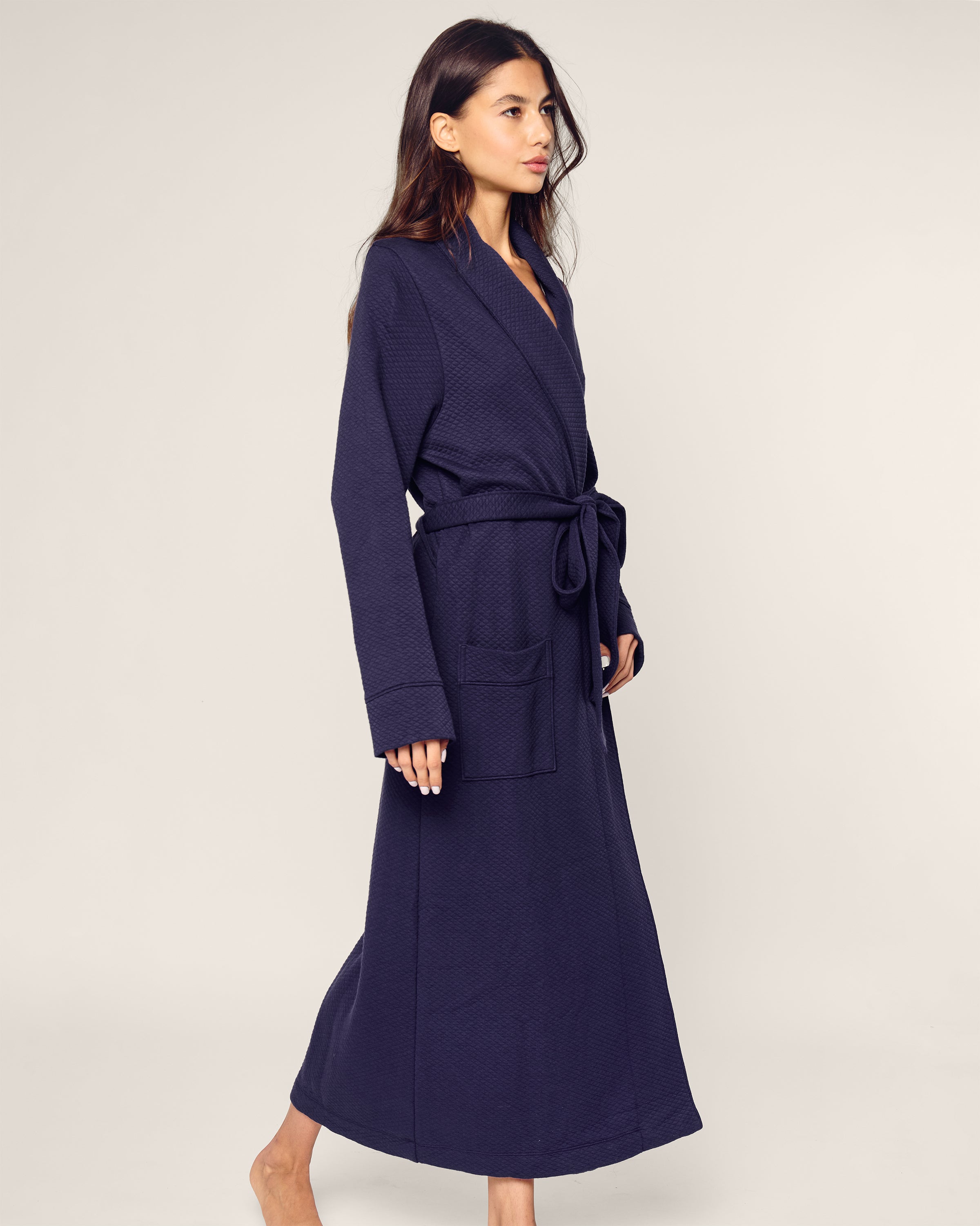 A person with long hair wears a luxury Womens Pima Ophelia Robe in Blue Dusk Jacquard by Petite Plume, tied at the waist. Made from Peruvian Pima cotton, it adds elegance as they walk barefoot against a plain background, looking to the side.