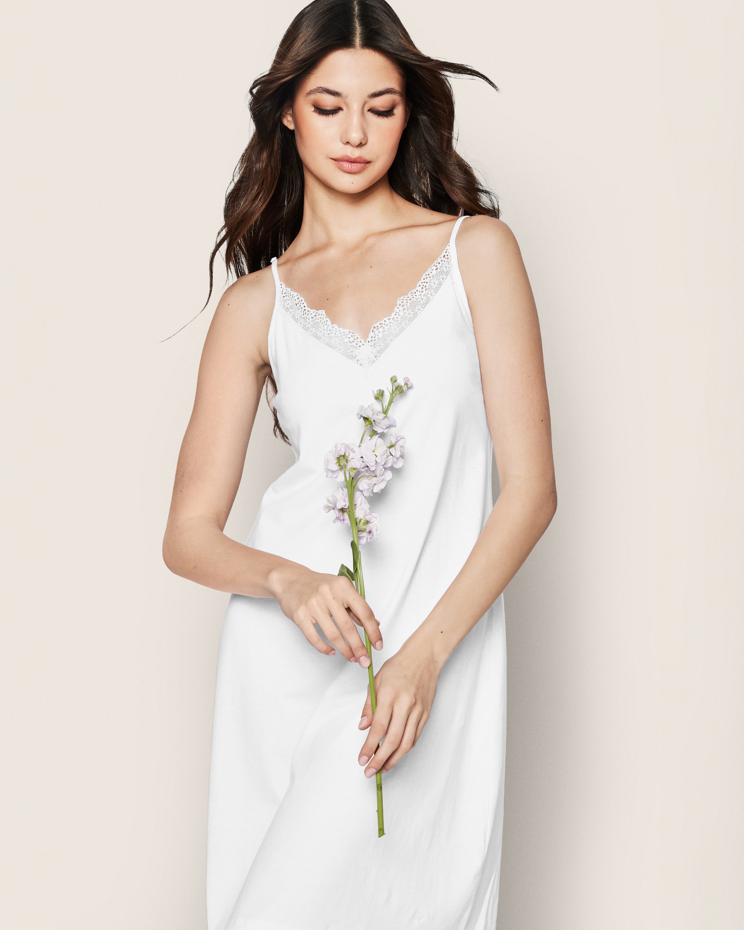 A woman wearing Petite Plumes Womens Pima Nightgown with Lace in White, made from soft Peruvian Pima cotton, holds a small bouquet of white flowers. She has long, wavy brown hair and poses against a plain, light-colored background.