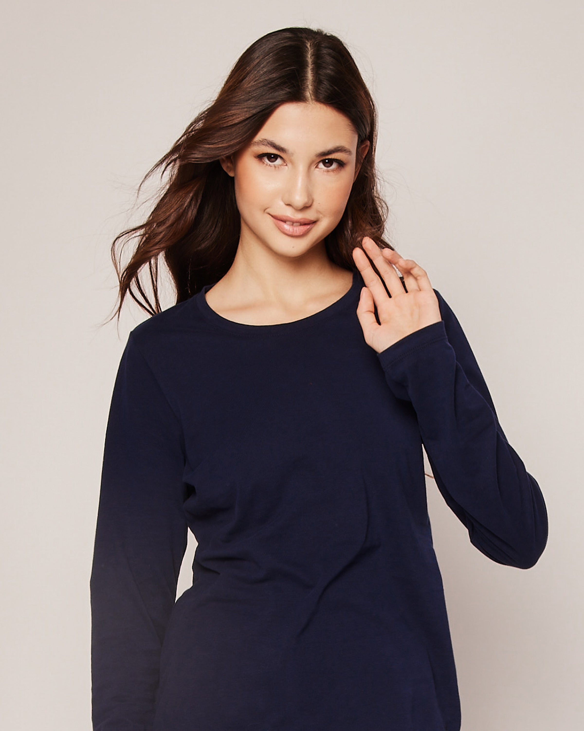 A woman with long brown hair wears a Petite Plume Womens Pima Long Sleeve Top in Navy, posing against a plain background while smiling gently and raising her right hand slightly.