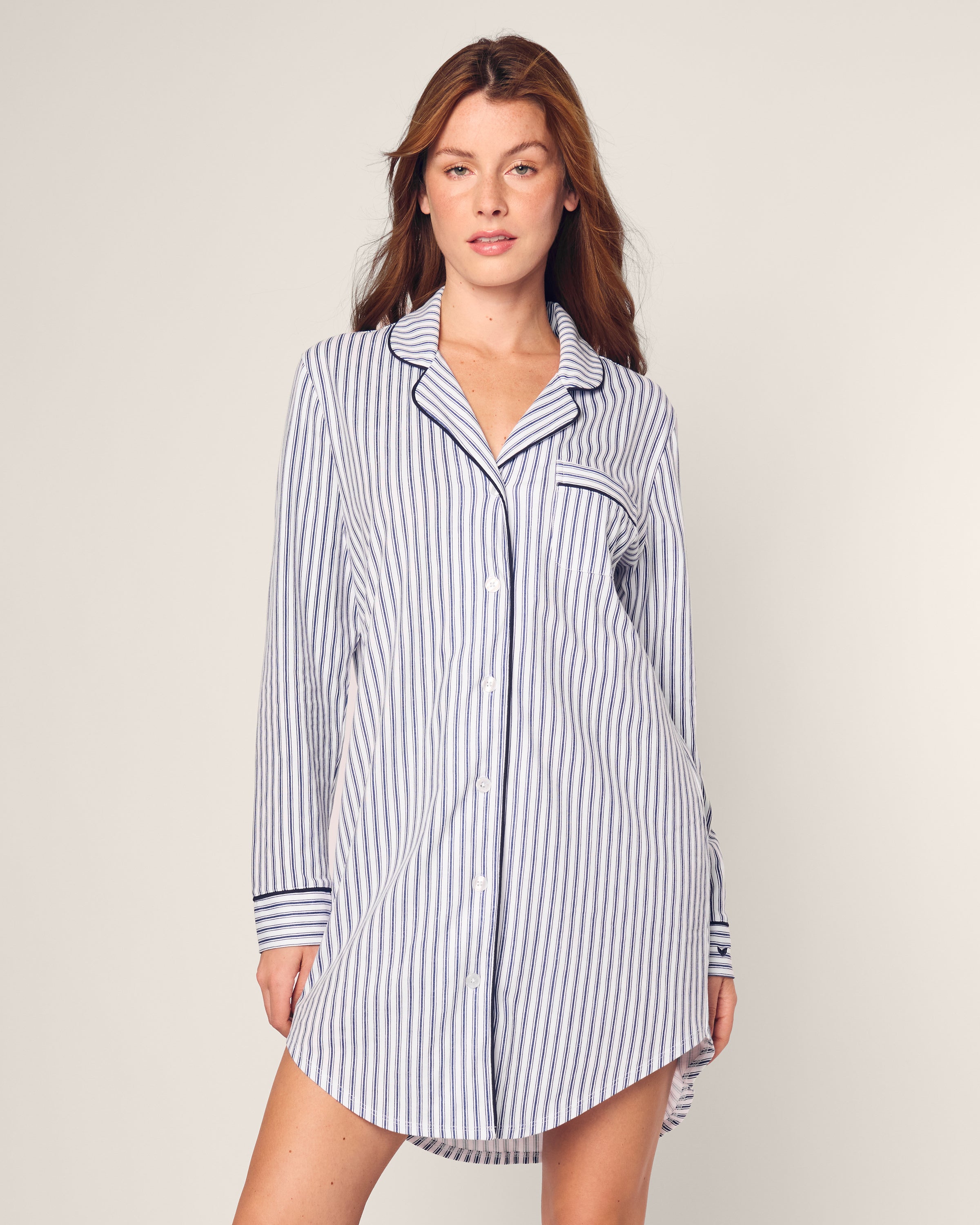 A person wearing the Womens Pima Nightshirt in Navy French Ticking by Petite Plume, made from exquisite Peruvian Pima cotton with blue and white stripes, stands against a light background with their hands relaxed at their sides.