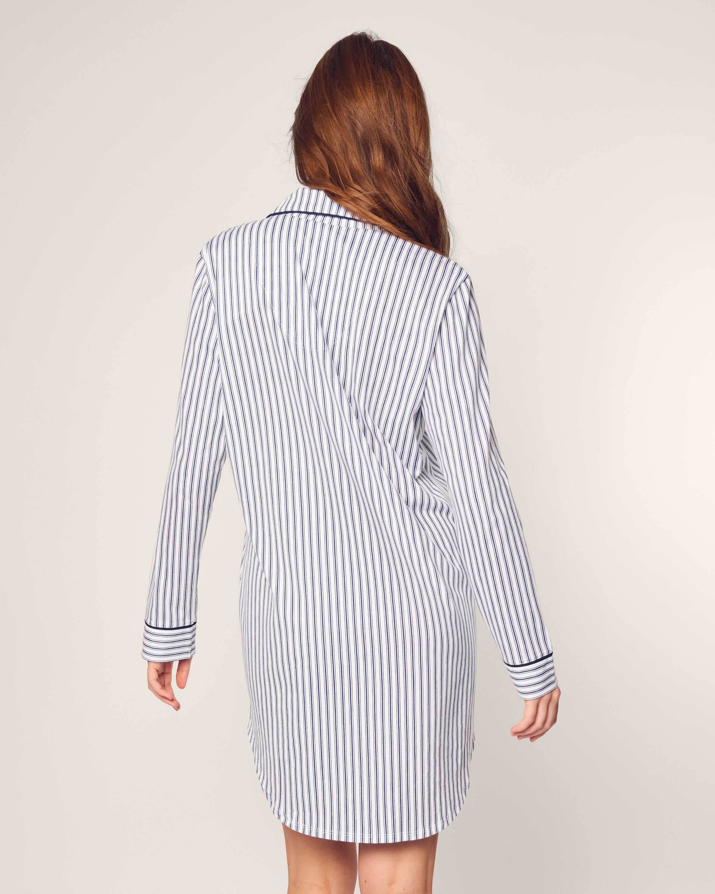 Against a plain background, a person with long hair wears the Womens Pima Nightshirt in Navy French Ticking by Petite Plume. It features long sleeves and a collar, combining the classic look of blue and white stripes with the comfort of luxury pajamas.