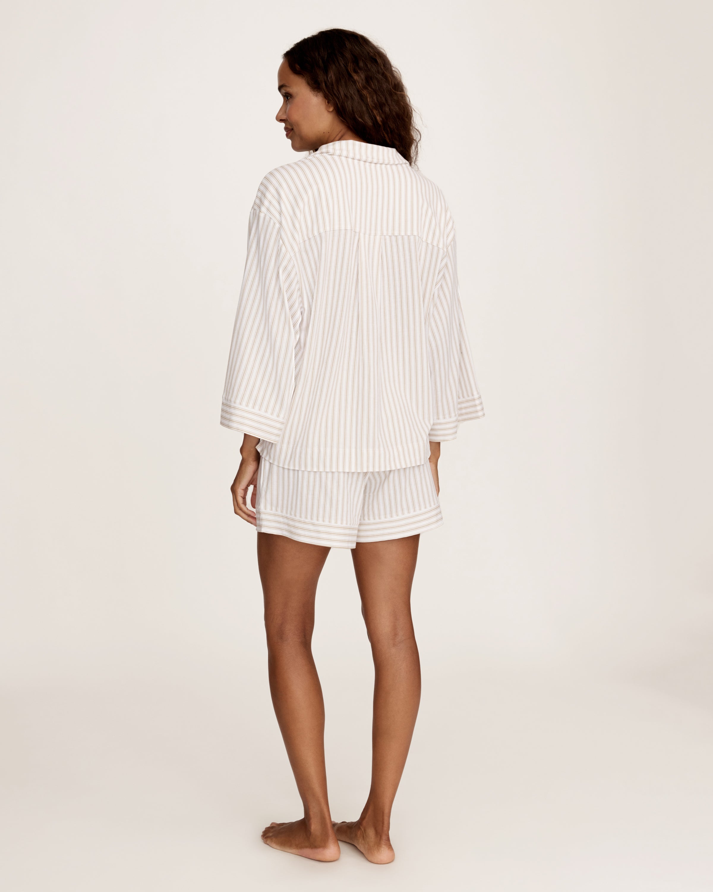 The Olivia Pima Short Set in Camel Ticking features a two-piece pajama set with beige and white vertical stripes, including a button-up, collared long-sleeve shirt with a breast pocket, and matching relaxed-fit drawstring shorts.