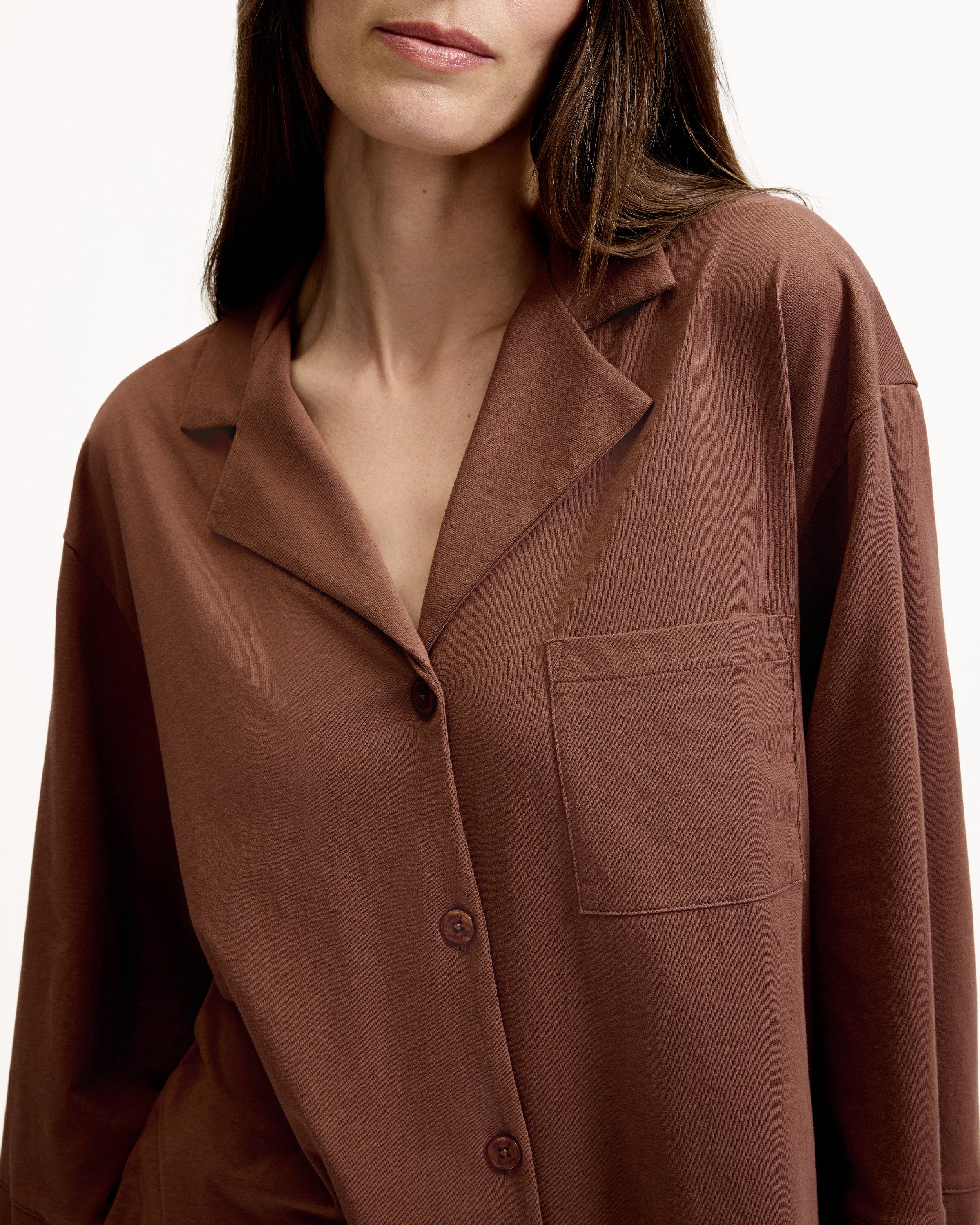 A woman stands barefoot against a white background, wearing The Olivia Lightweight Pima Short Set in Chocolate Brown—featuring a loose-fitting, long-sleeved button-up top with a pocket and matching shorts.