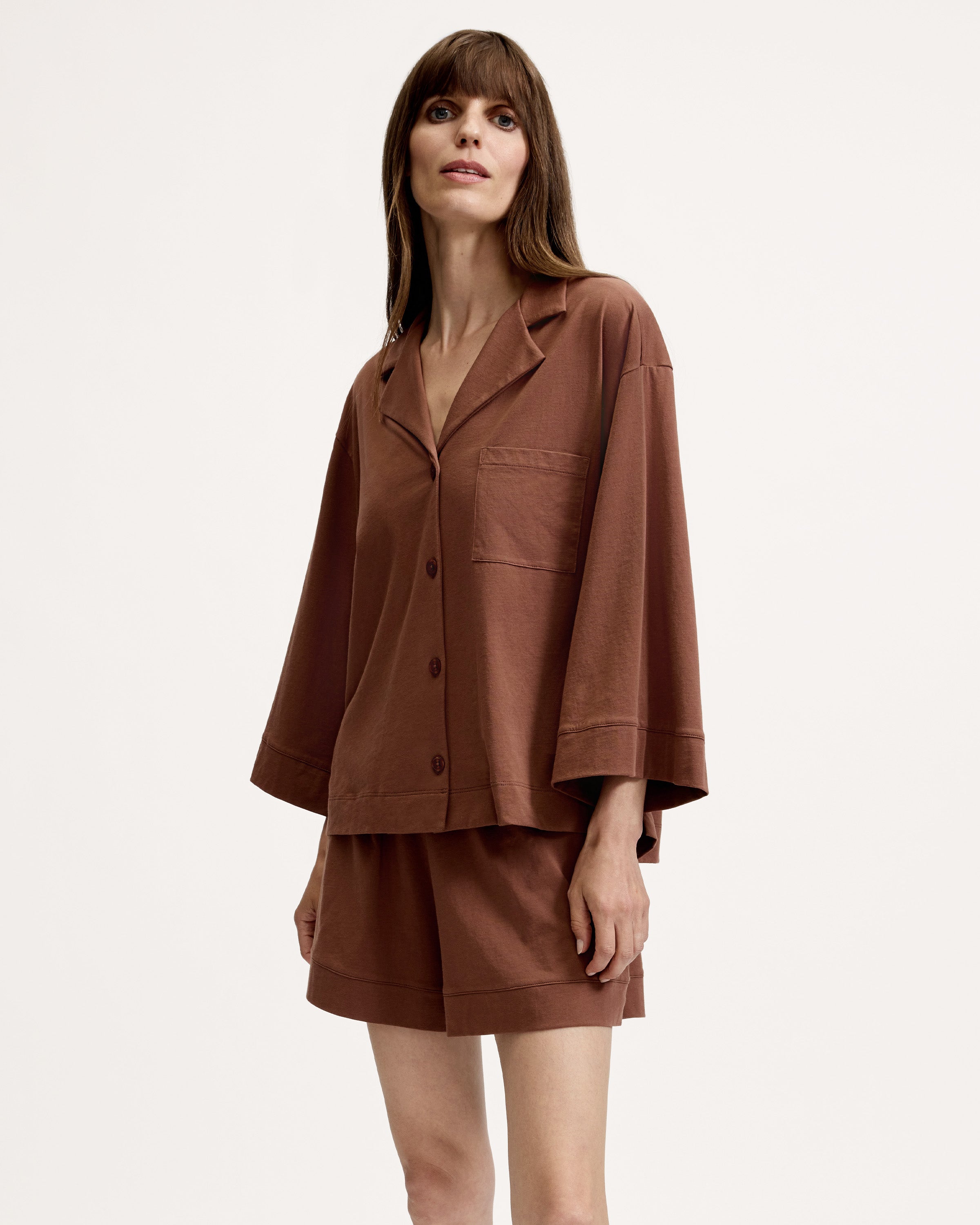 A woman stands barefoot against a white background, wearing The Olivia Lightweight Pima Short Set in Chocolate Brown—featuring a loose-fitting, long-sleeved button-up top with a pocket and matching shorts.