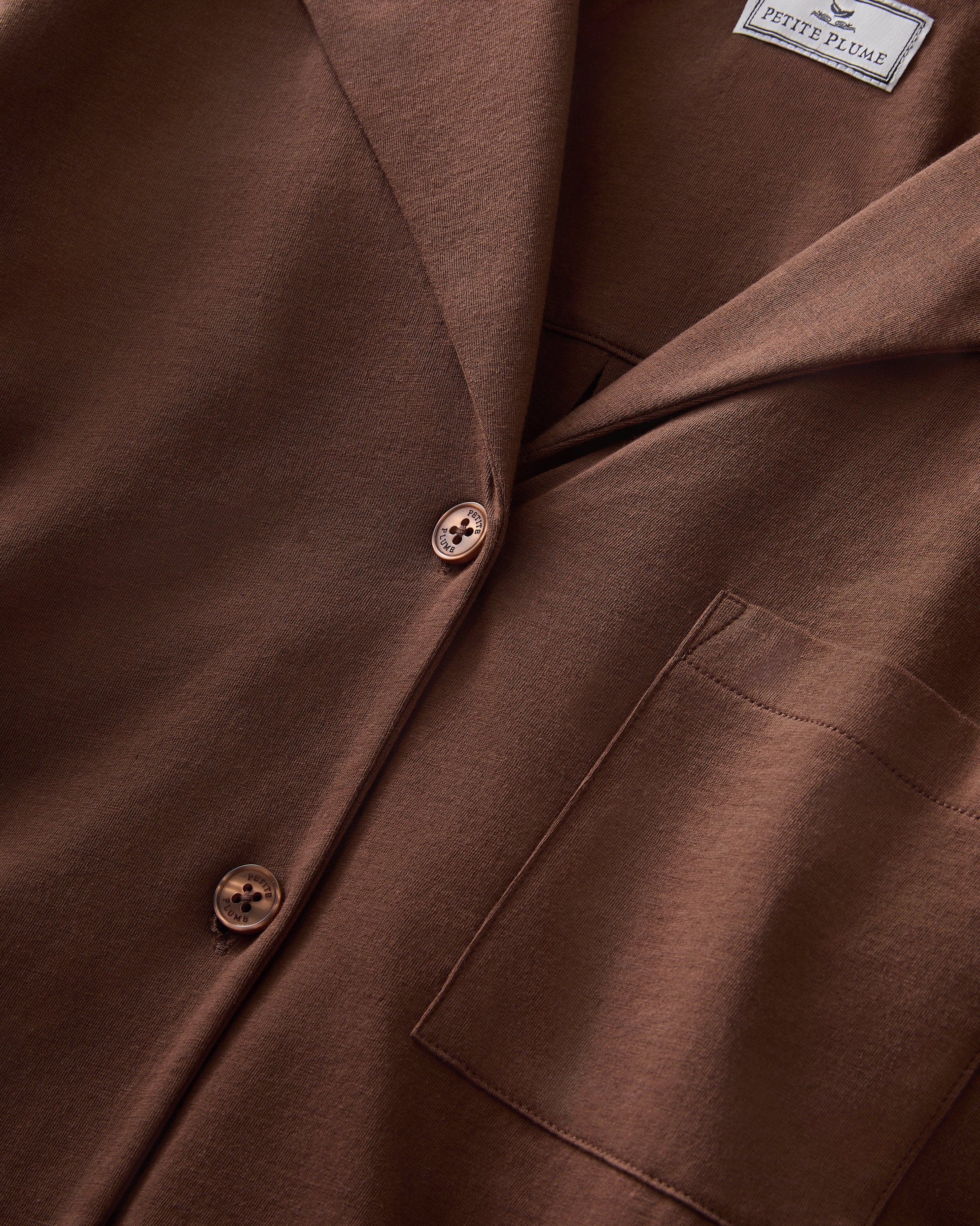 A woman stands barefoot against a white background, wearing The Olivia Lightweight Pima Short Set in Chocolate Brown—featuring a loose-fitting, long-sleeved button-up top with a pocket and matching shorts.