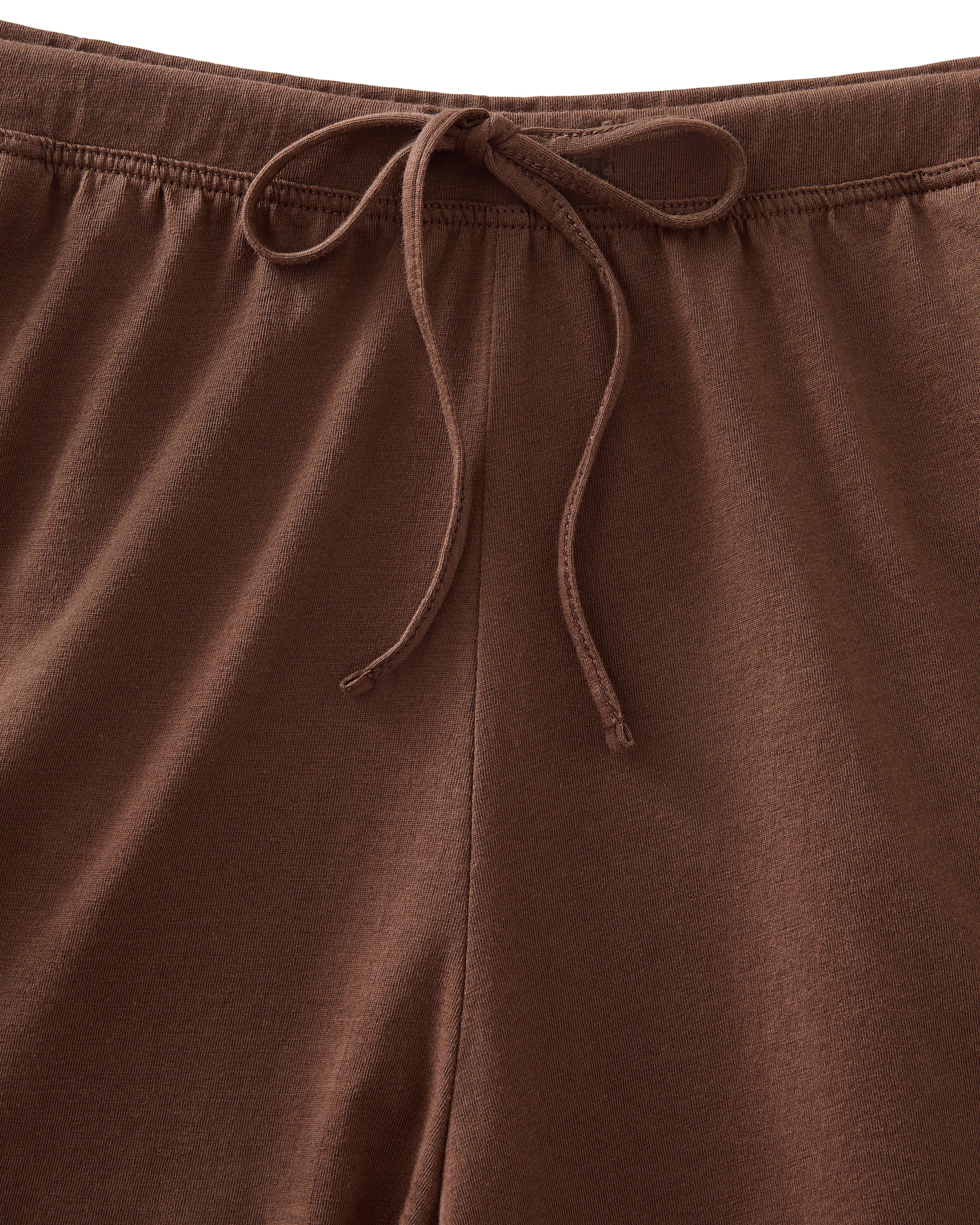 A woman stands barefoot against a white background, wearing The Olivia Lightweight Pima Short Set in Chocolate Brown—featuring a loose-fitting, long-sleeved button-up top with a pocket and matching shorts.