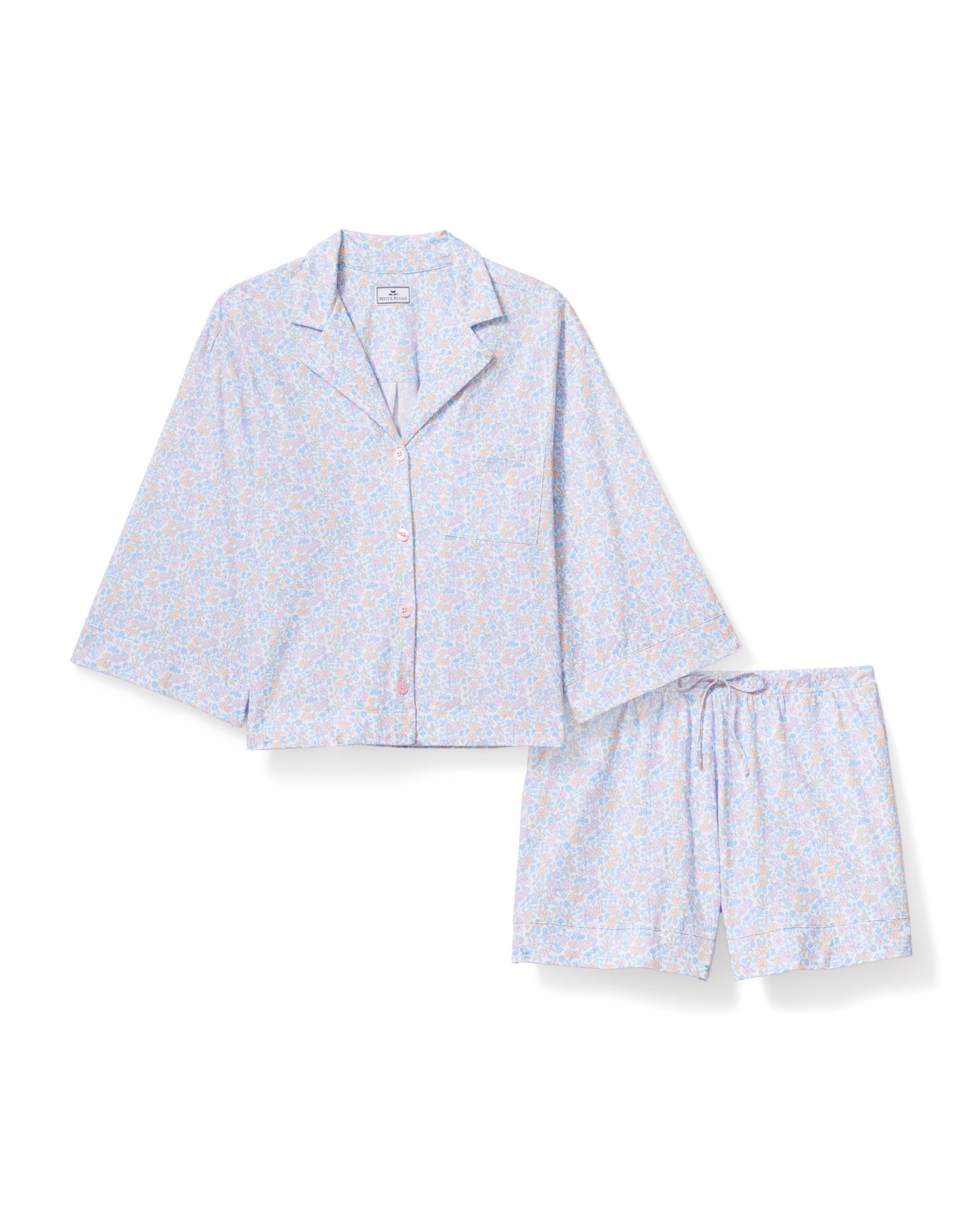 A woman with long, wavy hair wears The Olivia Lightweight Pima Short Set in Fleur Blanche, featuring a loose button-up top with wide sleeves and matching shorts adorned with a subtle floral pattern. She stands against a plain, light background.