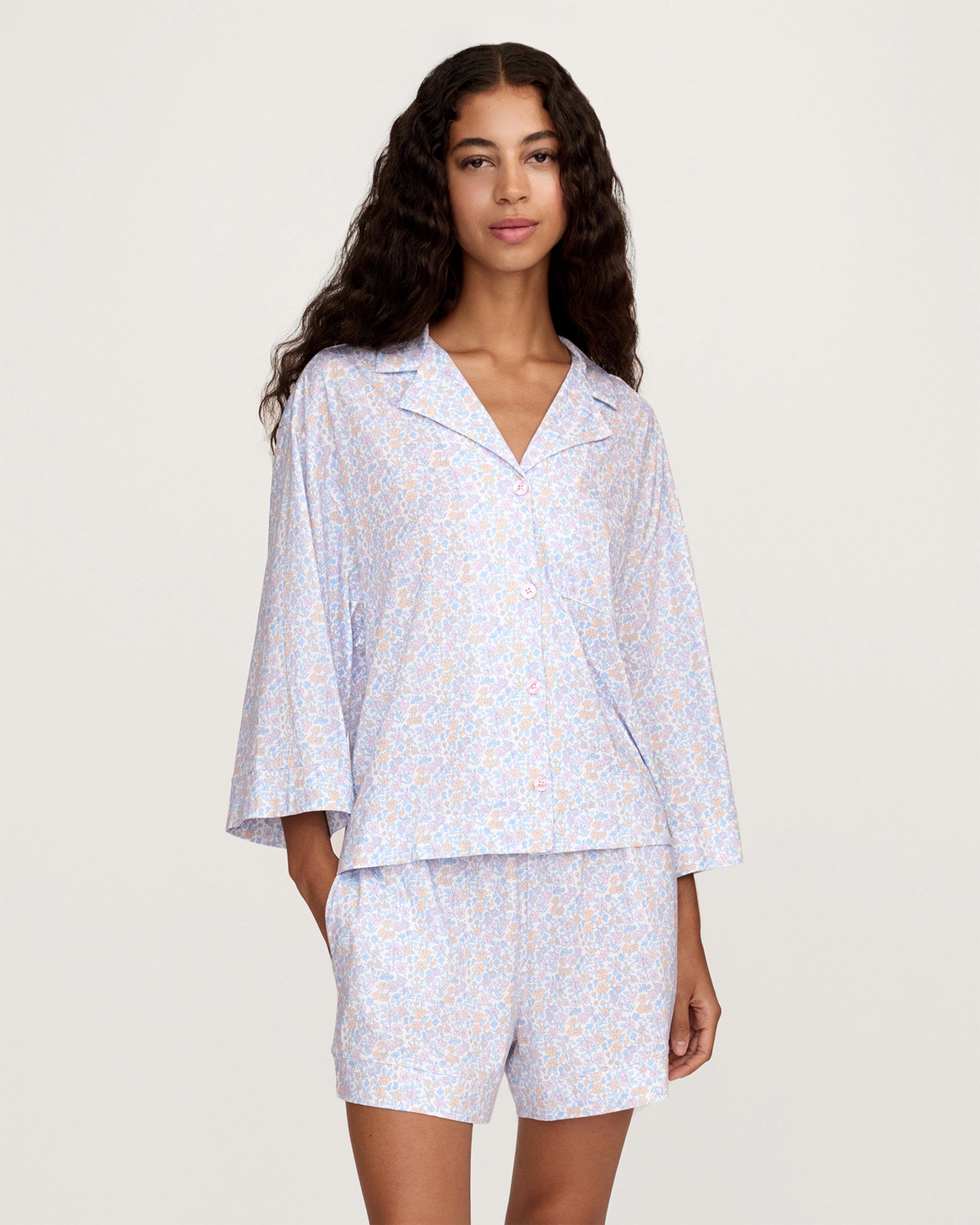 A woman with long, wavy hair wears The Olivia Lightweight Pima Short Set in Fleur Blanche, featuring a loose button-up top with wide sleeves and matching shorts adorned with a subtle floral pattern. She stands against a plain, light background.