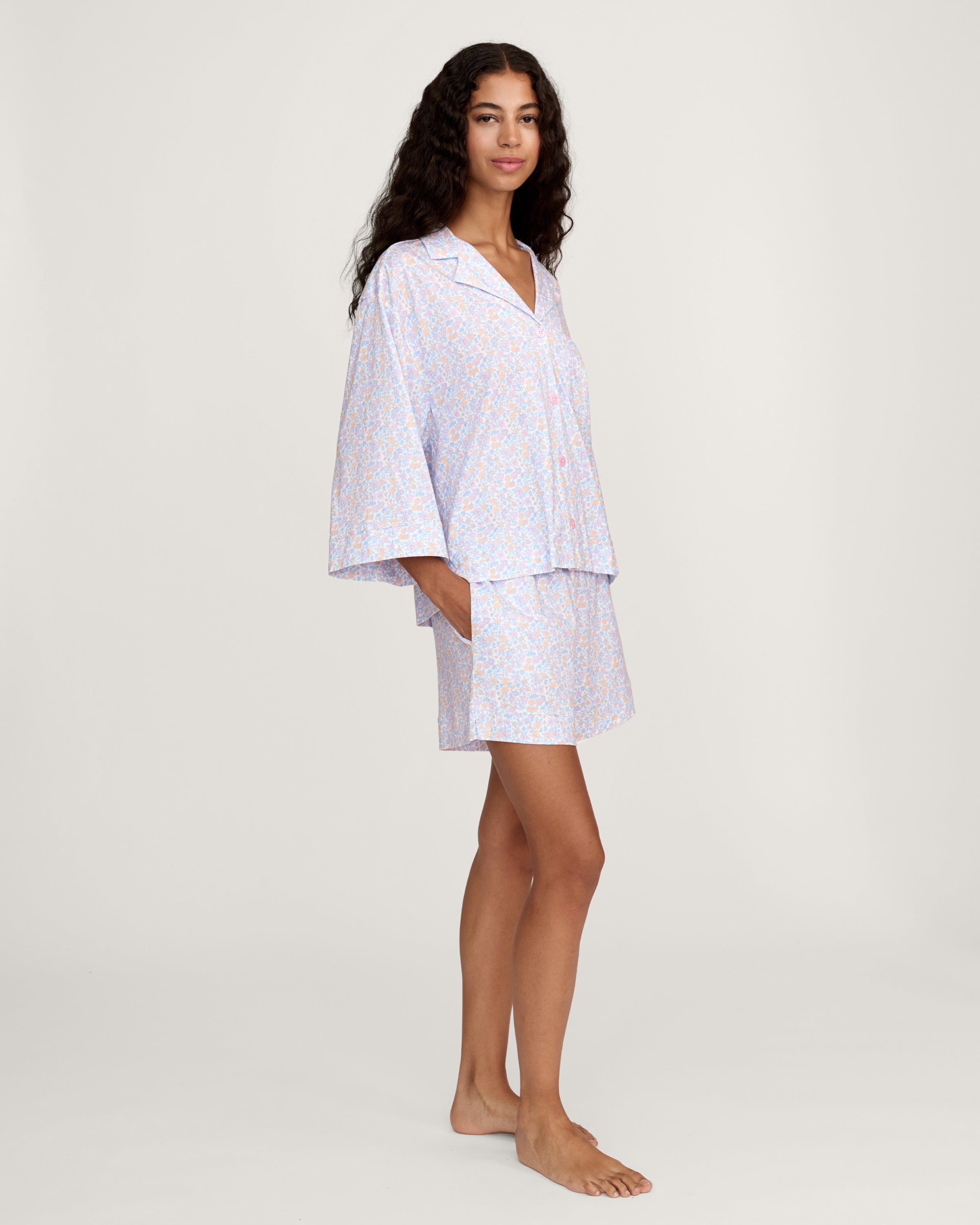 A woman with long, wavy hair wears The Olivia Lightweight Pima Short Set in Fleur Blanche, featuring a loose button-up top with wide sleeves and matching shorts adorned with a subtle floral pattern. She stands against a plain, light background.