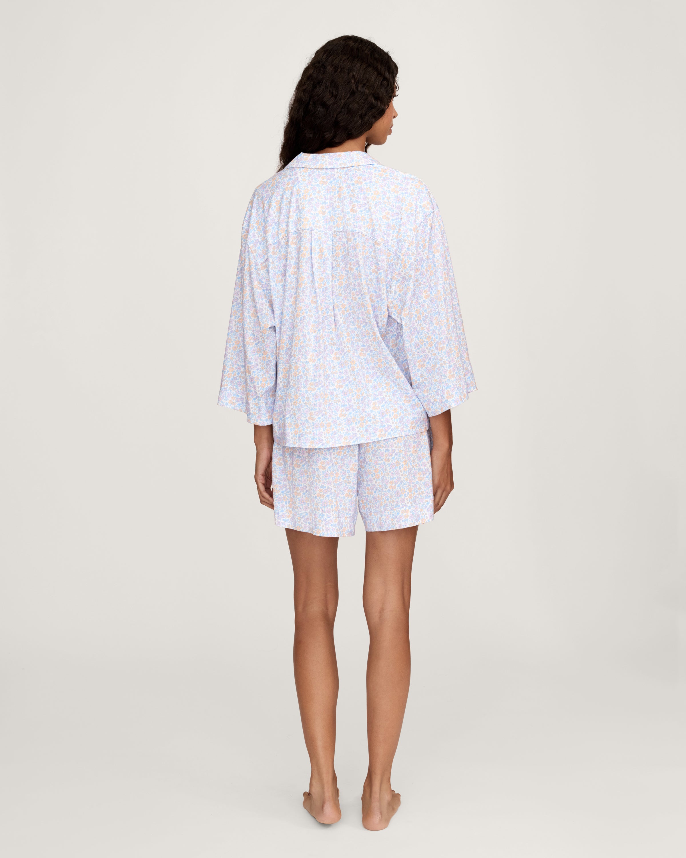 A woman with long, wavy hair wears The Olivia Lightweight Pima Short Set in Fleur Blanche, featuring a loose button-up top with wide sleeves and matching shorts adorned with a subtle floral pattern. She stands against a plain, light background.