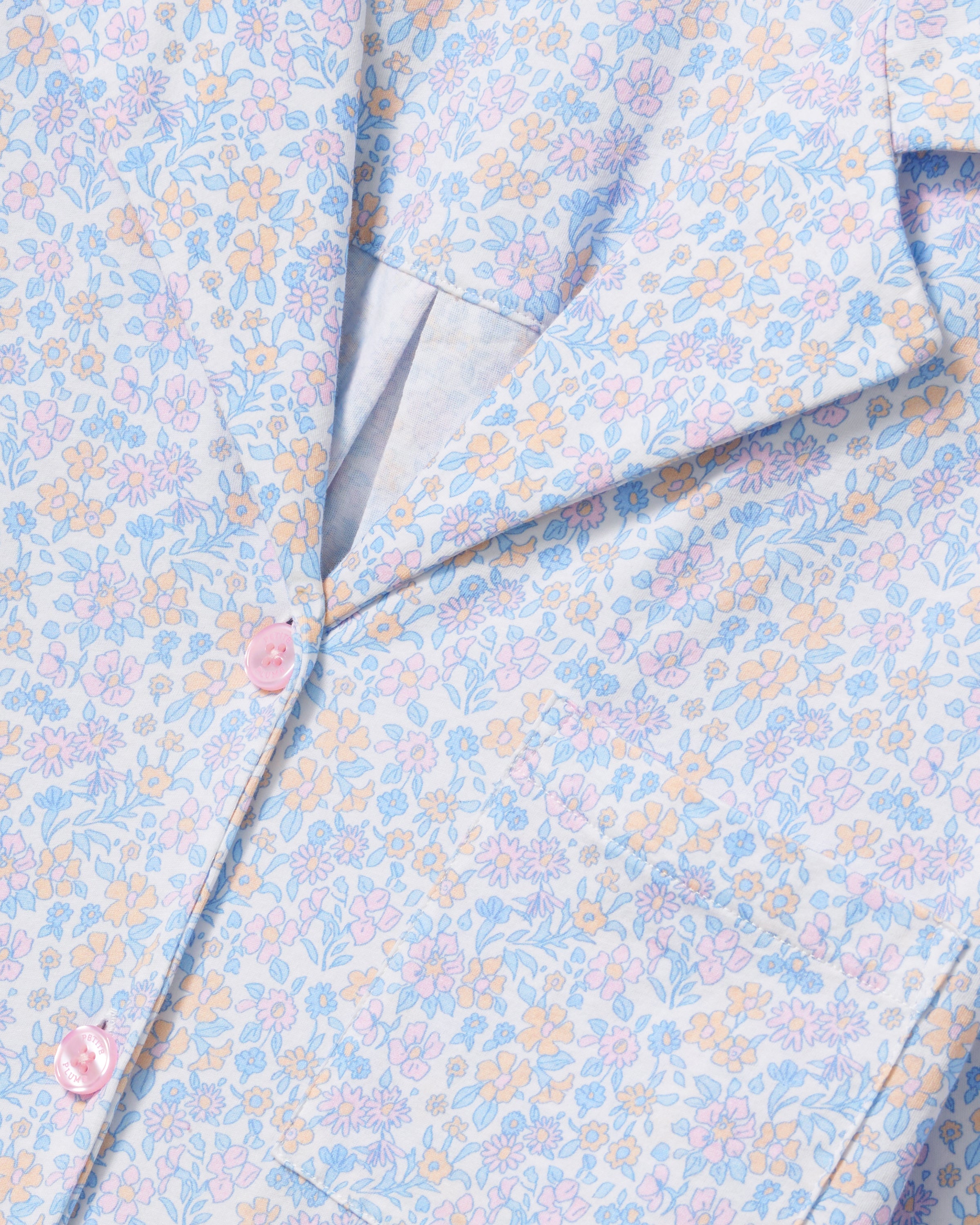 A woman with long, wavy hair wears The Olivia Lightweight Pima Short Set in Fleur Blanche, featuring a loose button-up top with wide sleeves and matching shorts adorned with a subtle floral pattern. She stands against a plain, light background.