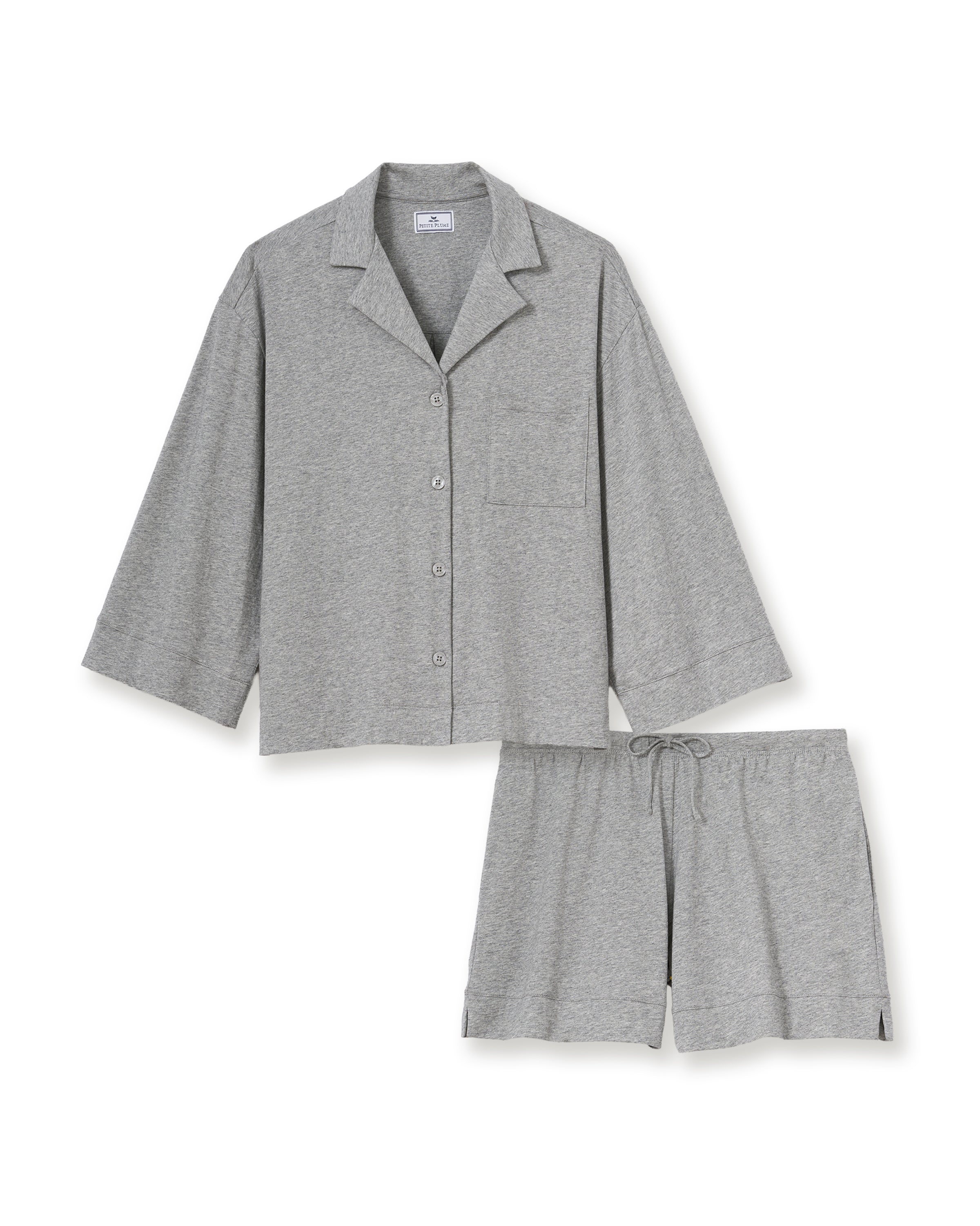 A woman with long, wavy hair and a neutral expression stands barefoot against a plain background, wearing The Olivia Lightweight Pima Short Set in Heather Grey—featuring a loose-fitting, long-sleeve top and matching shorts.