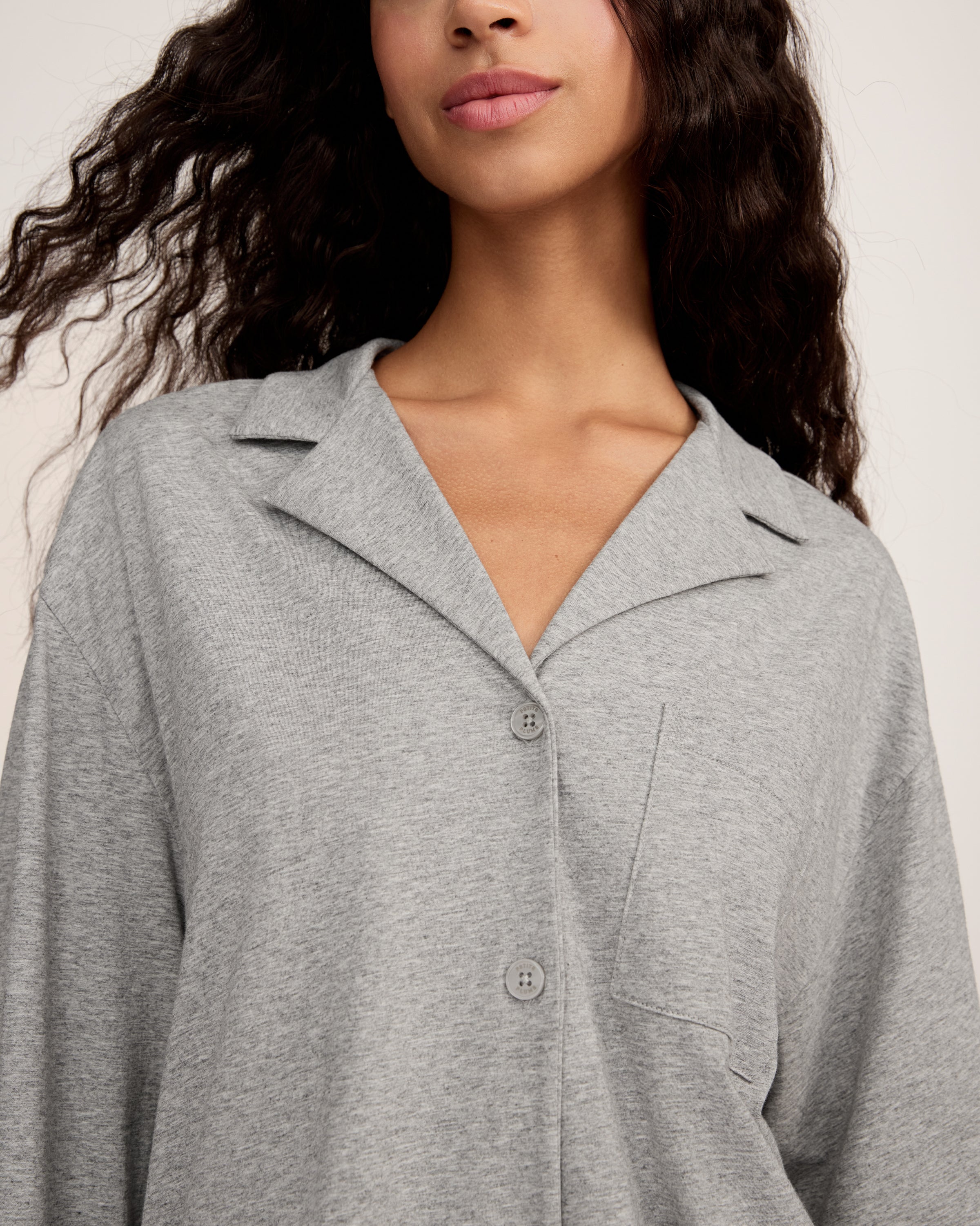 A woman with long, wavy hair and a neutral expression stands barefoot against a plain background, wearing The Olivia Lightweight Pima Short Set in Heather Grey—featuring a loose-fitting, long-sleeve top and matching shorts.