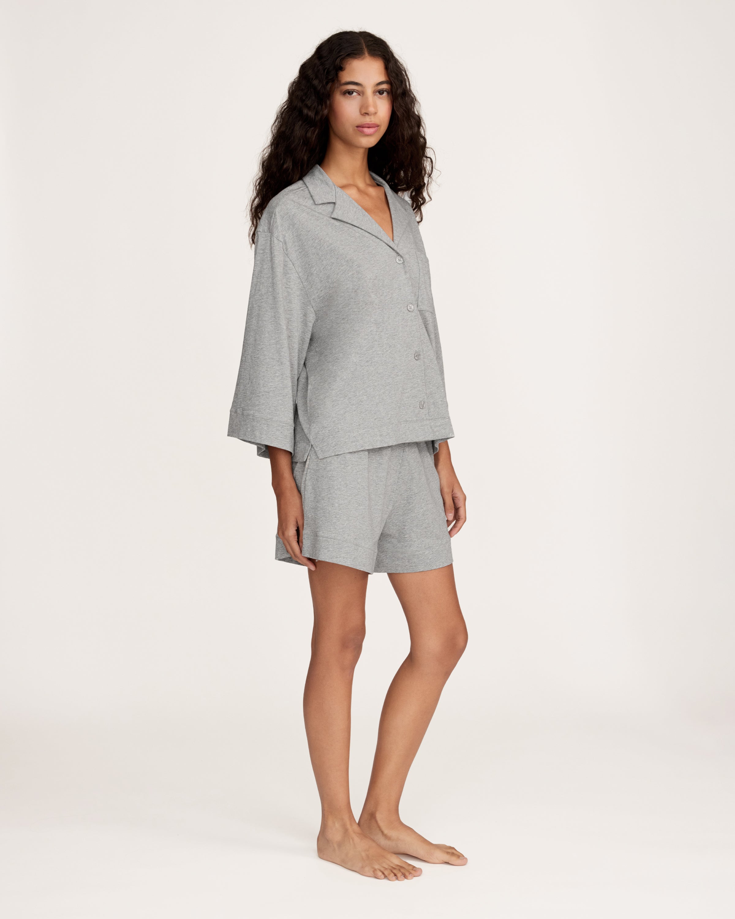 A woman with long, wavy hair and a neutral expression stands barefoot against a plain background, wearing The Olivia Lightweight Pima Short Set in Heather Grey—featuring a loose-fitting, long-sleeve top and matching shorts.