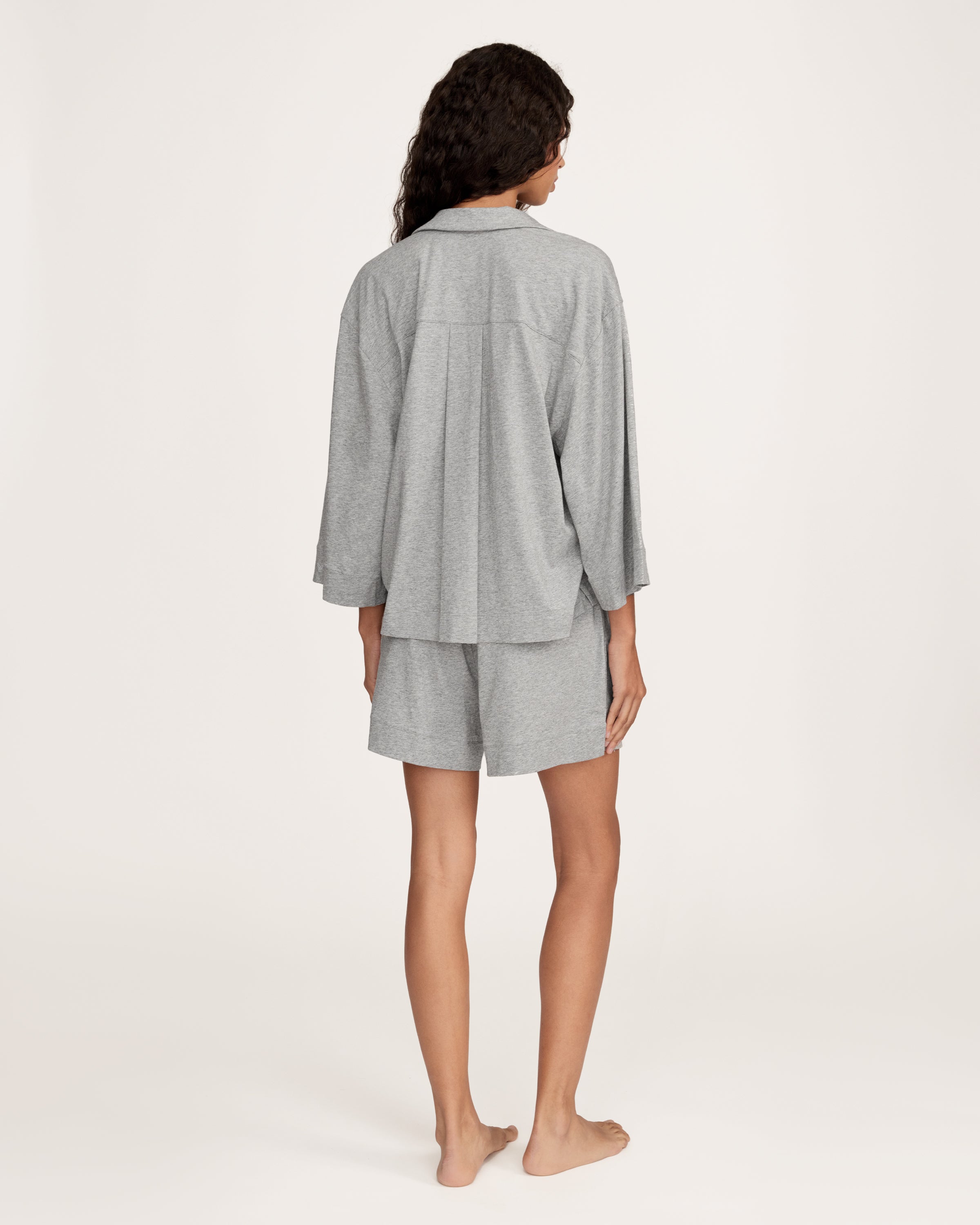 A woman with long, wavy hair and a neutral expression stands barefoot against a plain background, wearing The Olivia Lightweight Pima Short Set in Heather Grey—featuring a loose-fitting, long-sleeve top and matching shorts.