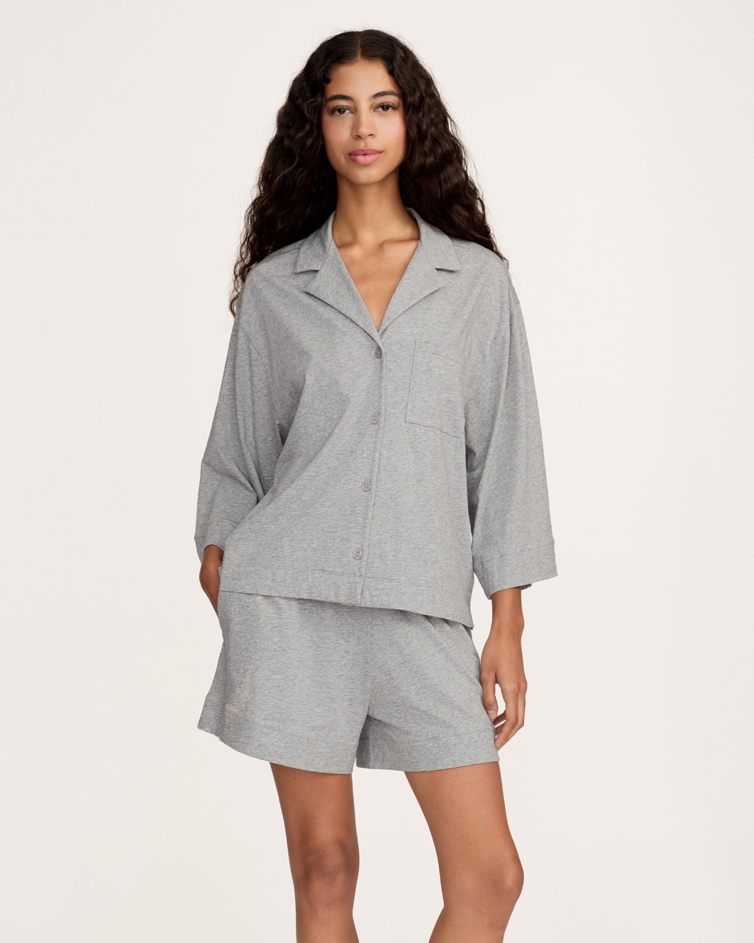 A woman with long, wavy hair and a neutral expression stands barefoot against a plain background, wearing The Olivia Lightweight Pima Short Set in Heather Grey—featuring a loose-fitting, long-sleeve top and matching shorts.