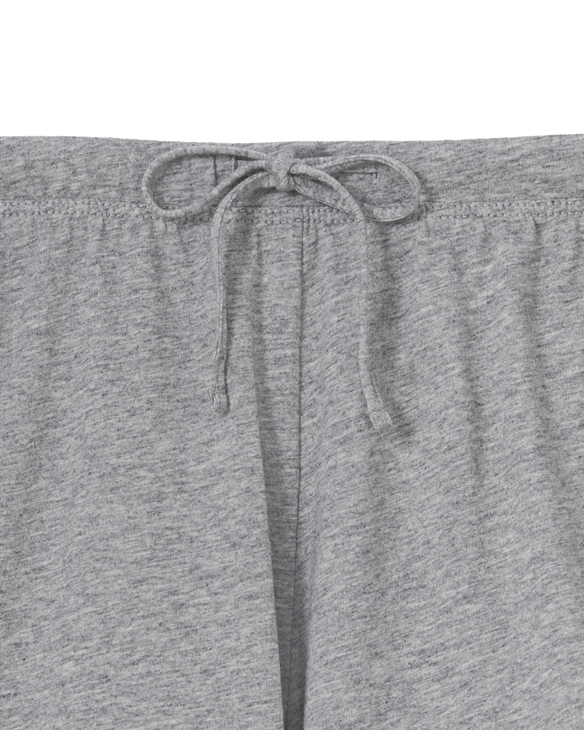 A woman with long, wavy hair and a neutral expression stands barefoot against a plain background, wearing The Olivia Lightweight Pima Short Set in Heather Grey—featuring a loose-fitting, long-sleeve top and matching shorts.