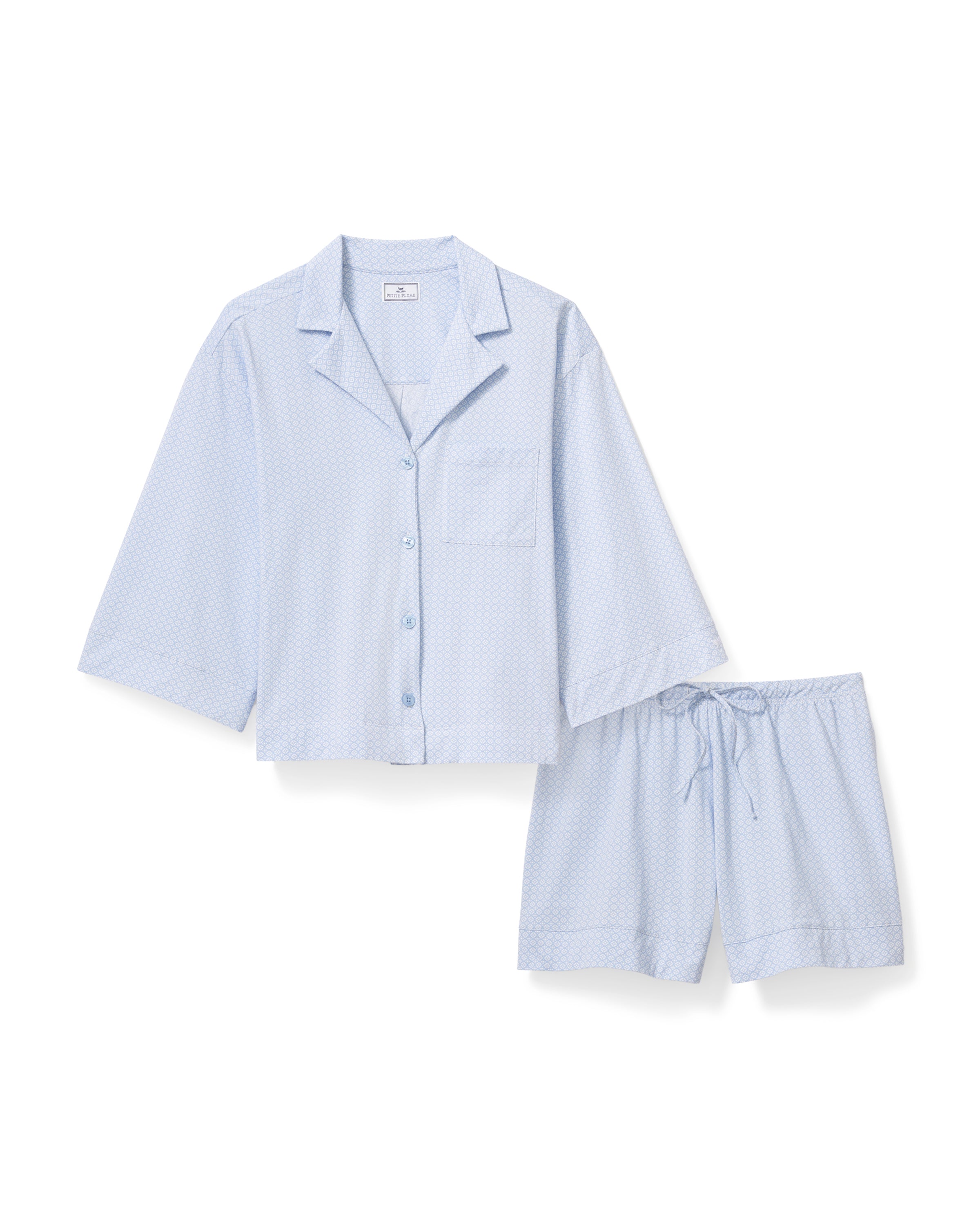 A woman with long curly hair models The Olivia Lightweight Pima Short Set in Periwinkle Geo—a light blue, long-sleeved button-up pajama top and matching dotted shorts—against a plain light background.