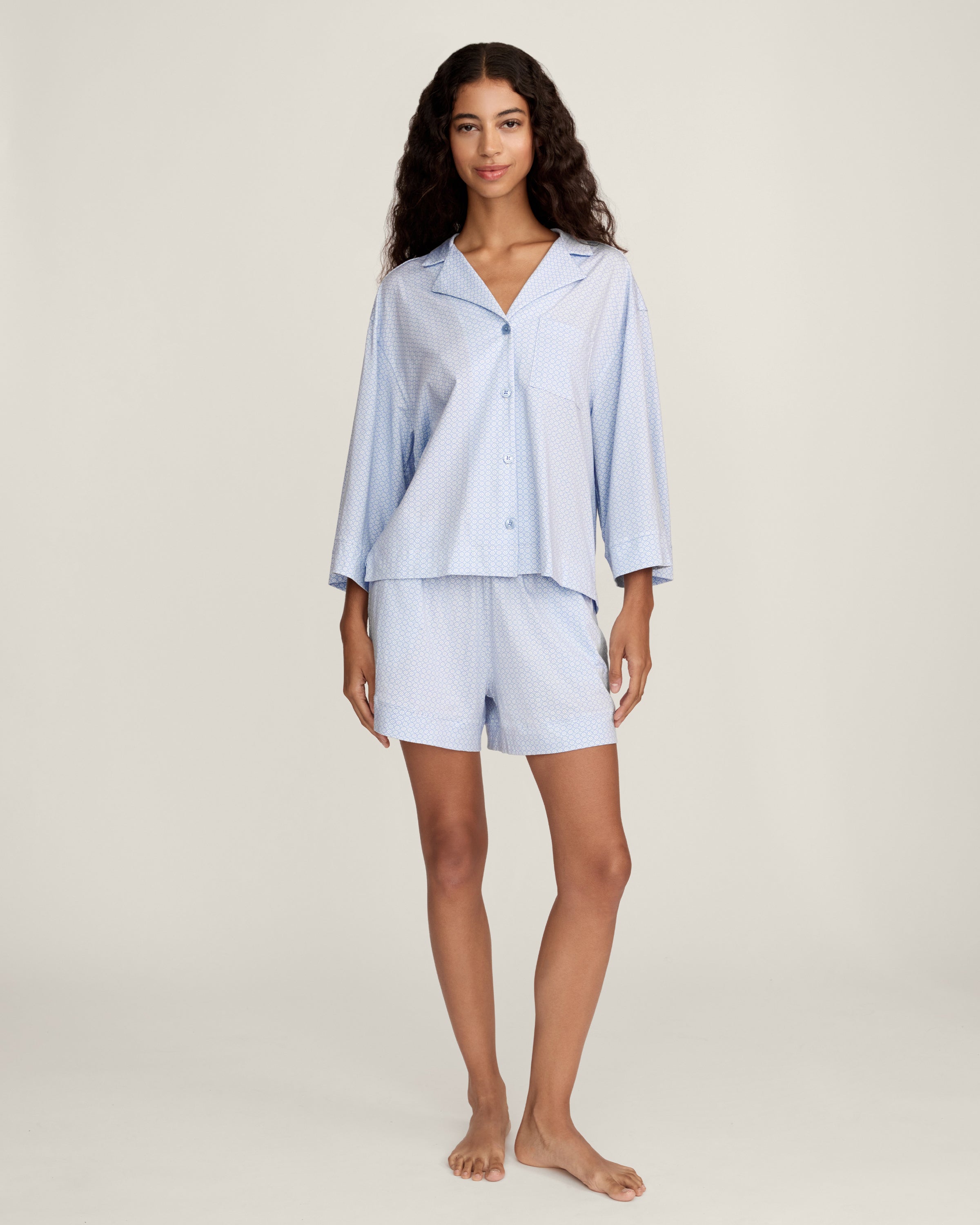 A woman with long curly hair models The Olivia Lightweight Pima Short Set in Periwinkle Geo—a light blue, long-sleeved button-up pajama top and matching dotted shorts—against a plain light background.