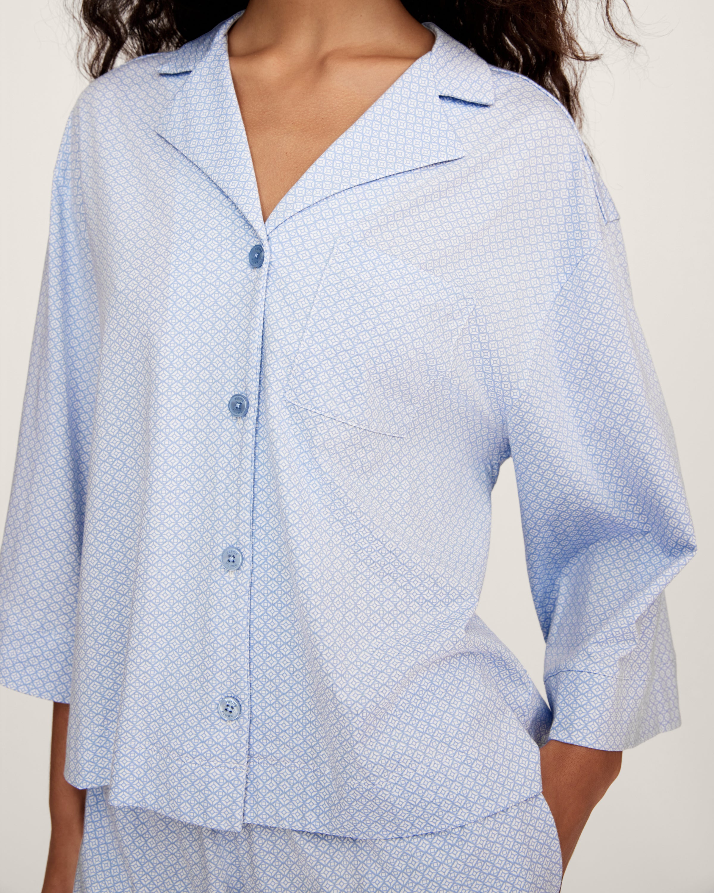 A woman with long curly hair models The Olivia Lightweight Pima Short Set in Periwinkle Geo—a light blue, long-sleeved button-up pajama top and matching dotted shorts—against a plain light background.