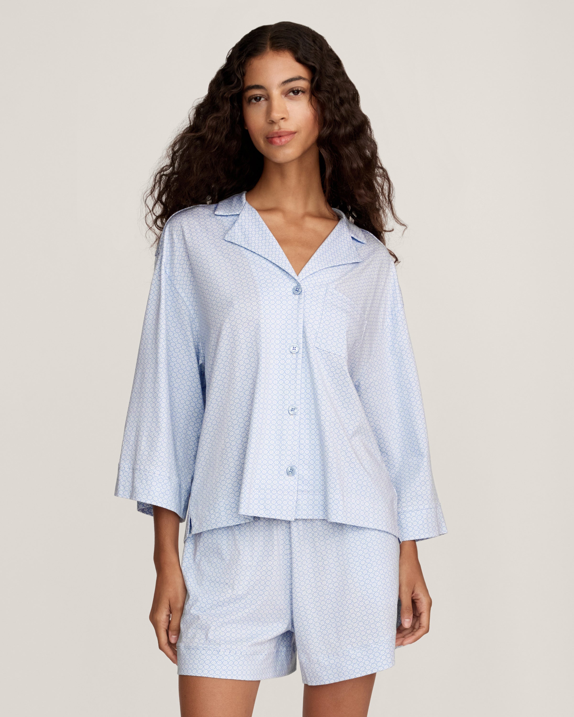 A woman with long curly hair models The Olivia Lightweight Pima Short Set in Periwinkle Geo—a light blue, long-sleeved button-up pajama top and matching dotted shorts—against a plain light background.
