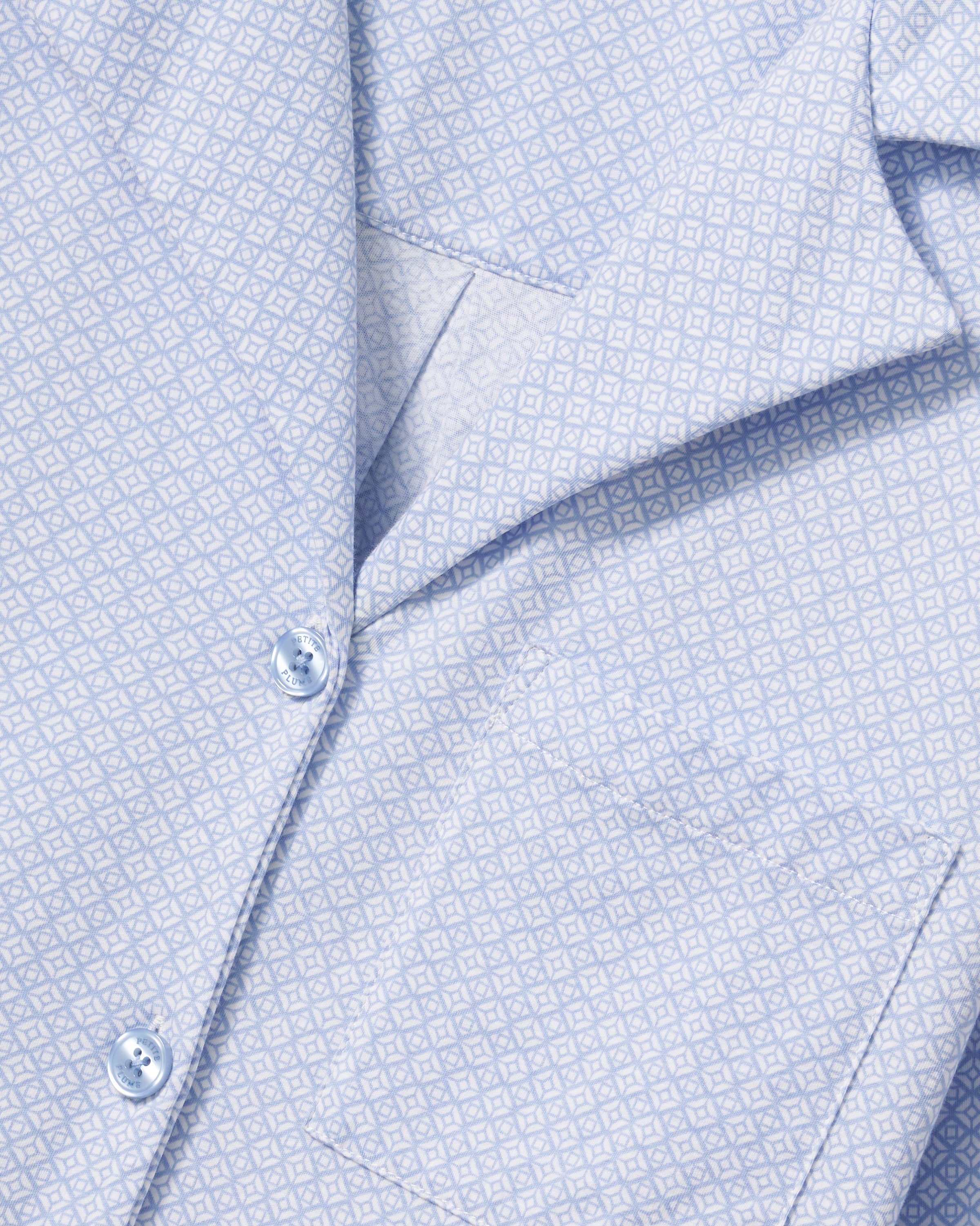 A woman with long curly hair models The Olivia Lightweight Pima Short Set in Periwinkle Geo—a light blue, long-sleeved button-up pajama top and matching dotted shorts—against a plain light background.