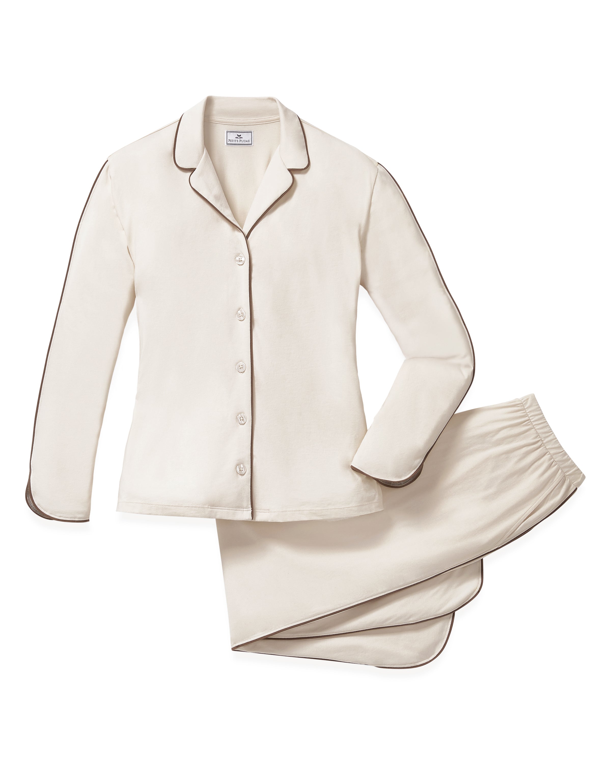 A person stands barefoot on a white background, wearing The Margaux Lightweight Pima Pajama Set in Ivory, featuring long sleeves, buttons, and pants with dark piping along the edges.