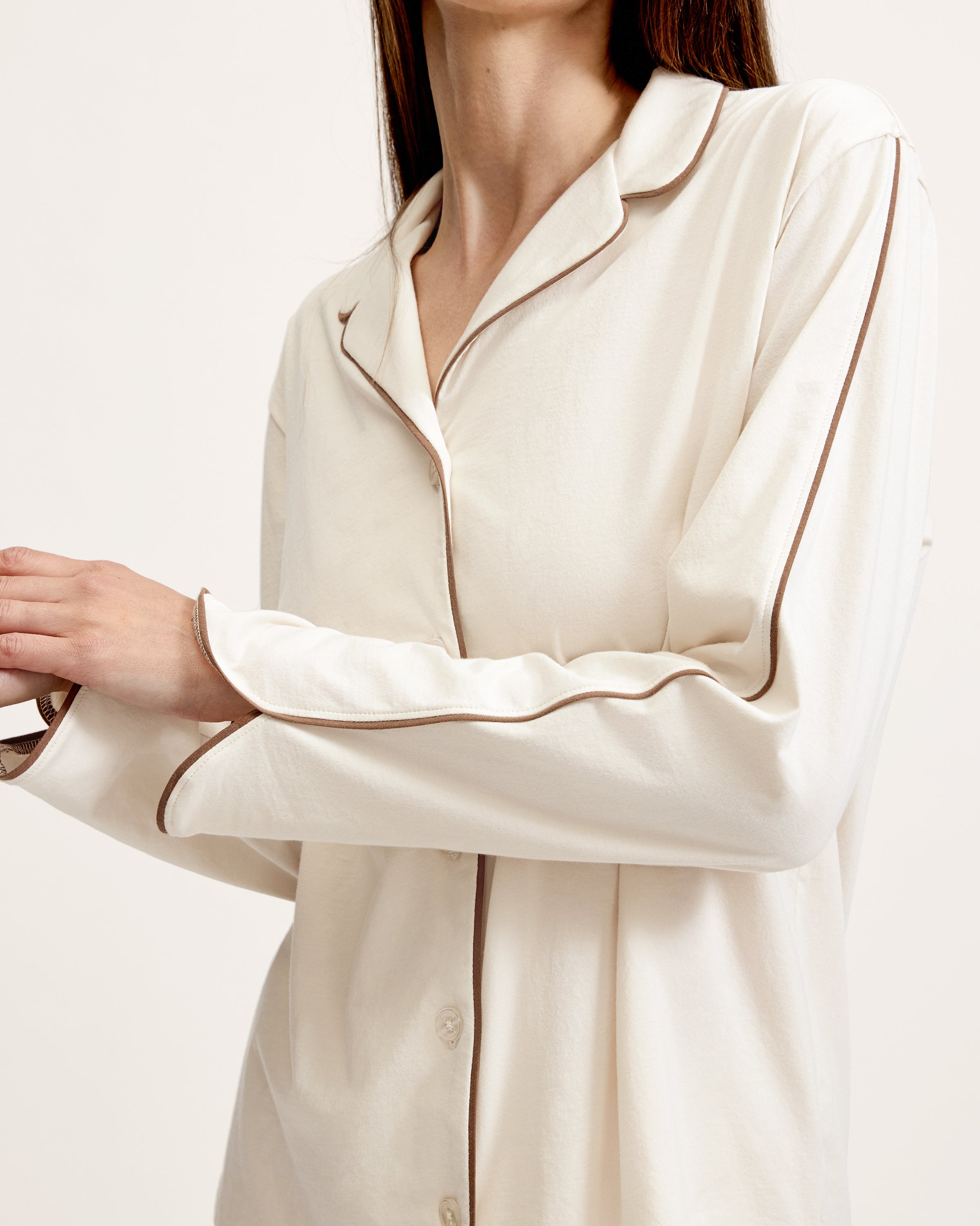 A person stands barefoot on a white background, wearing The Margaux Lightweight Pima Pajama Set in Ivory, featuring long sleeves, buttons, and pants with dark piping along the edges.