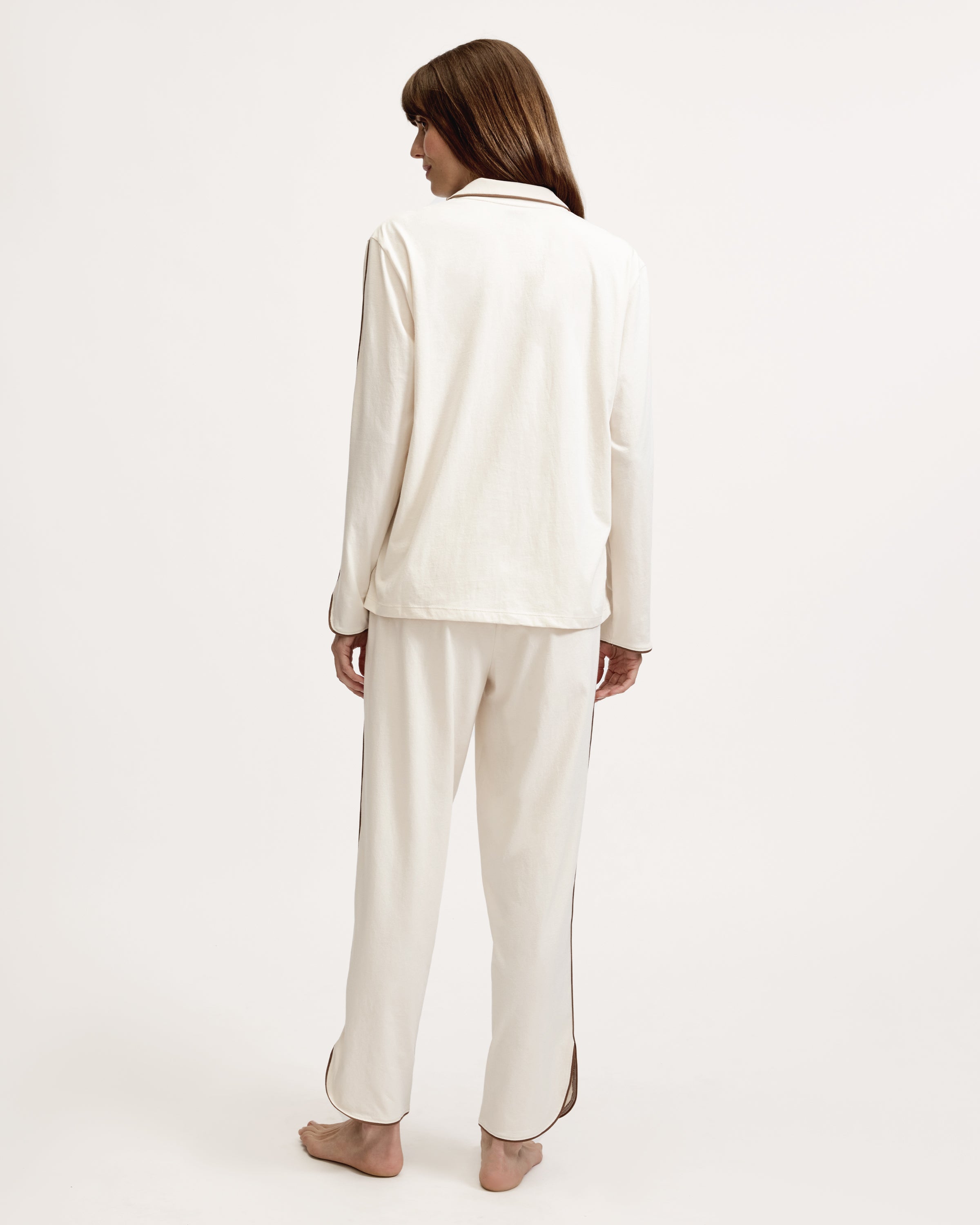 A person stands barefoot on a white background, wearing The Margaux Lightweight Pima Pajama Set in Ivory, featuring long sleeves, buttons, and pants with dark piping along the edges.