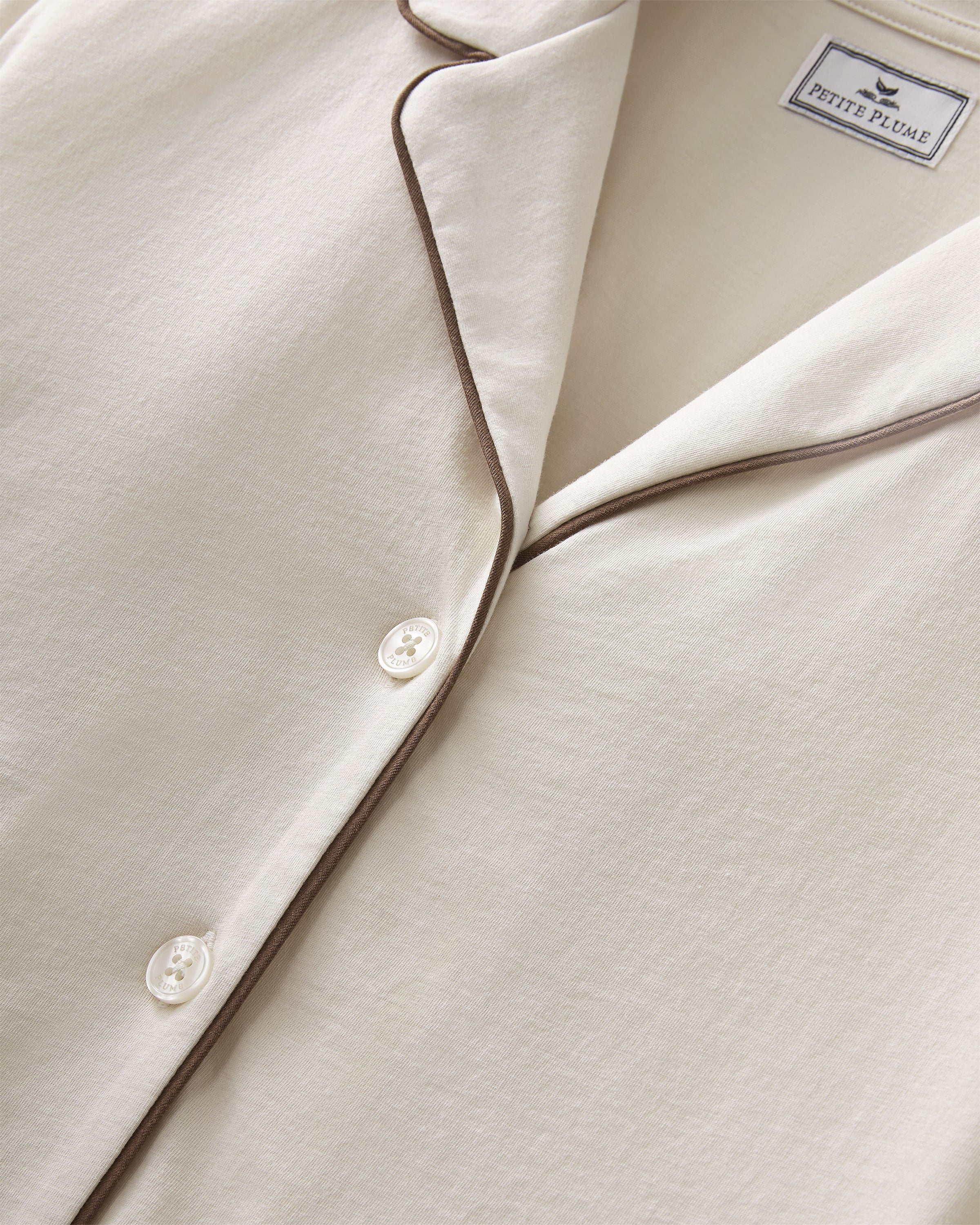 A person stands barefoot on a white background, wearing The Margaux Lightweight Pima Pajama Set in Ivory, featuring long sleeves, buttons, and pants with dark piping along the edges.