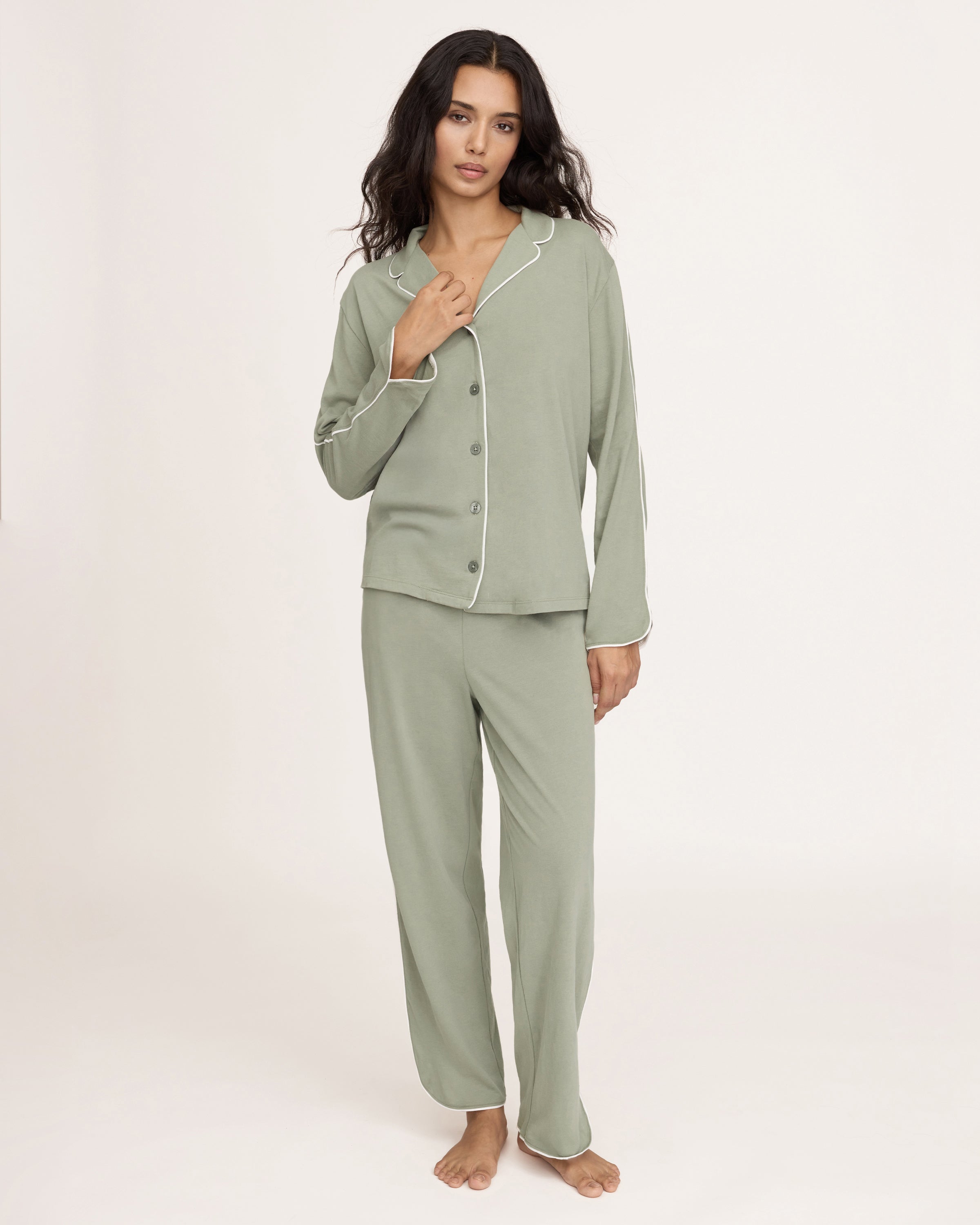 A woman with long dark hair wears The Margaux Pima Pajama Set in Sage, featuring a button-up long-sleeve top with white piping and matching pants. She stands barefoot against a plain, light background.
