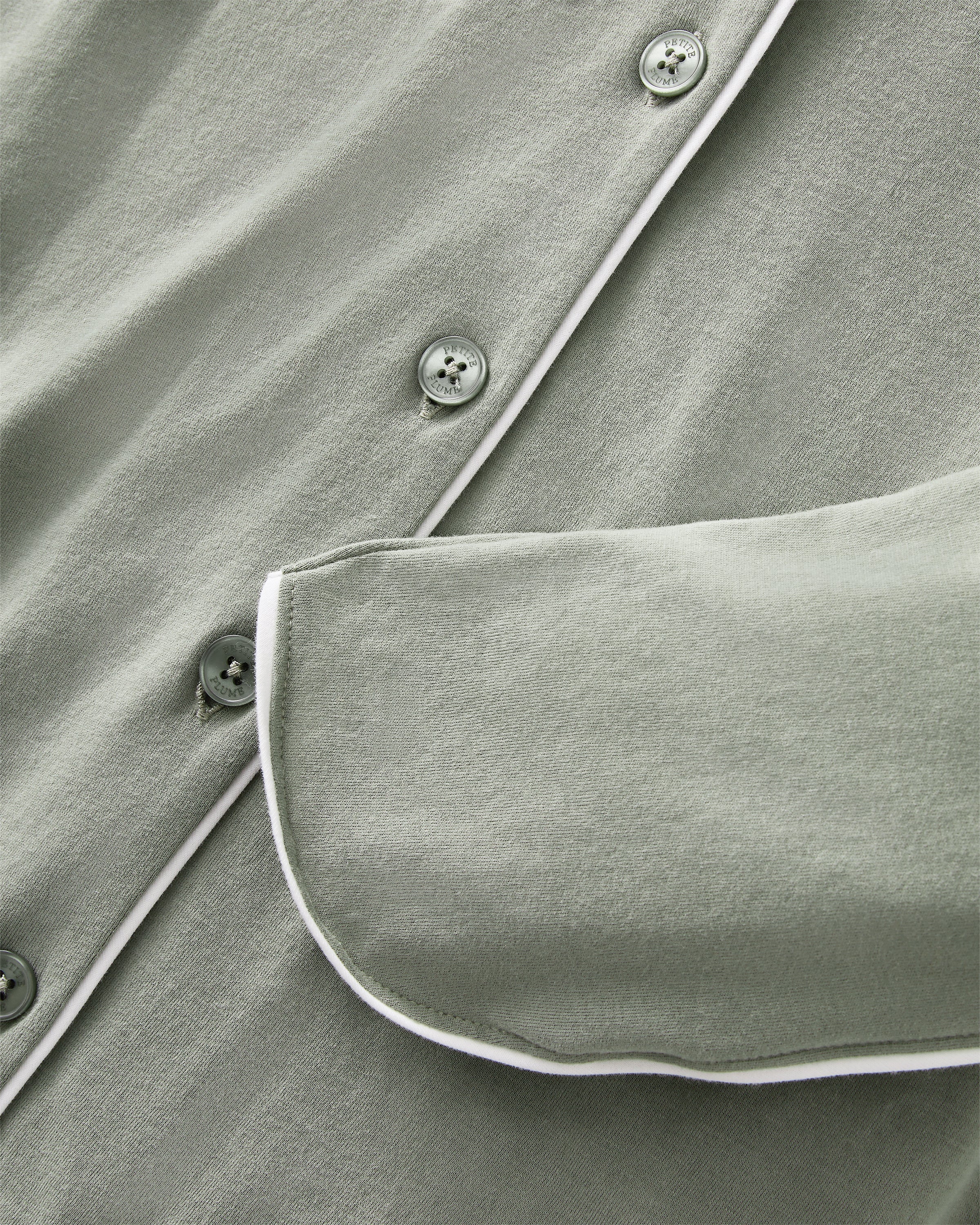 A woman with long dark hair wears The Margaux Pima Pajama Set in Sage, featuring a button-up long-sleeve top with white piping and matching pants. She stands barefoot against a plain, light background.