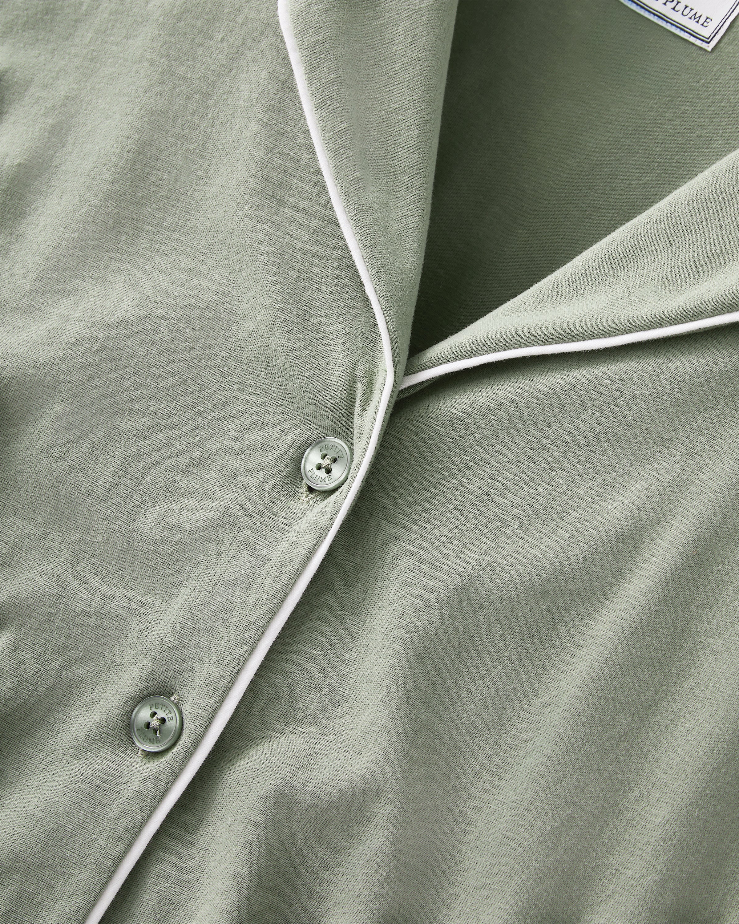 A woman with long dark hair wears The Margaux Pima Pajama Set in Sage, featuring a button-up long-sleeve top with white piping and matching pants. She stands barefoot against a plain, light background.