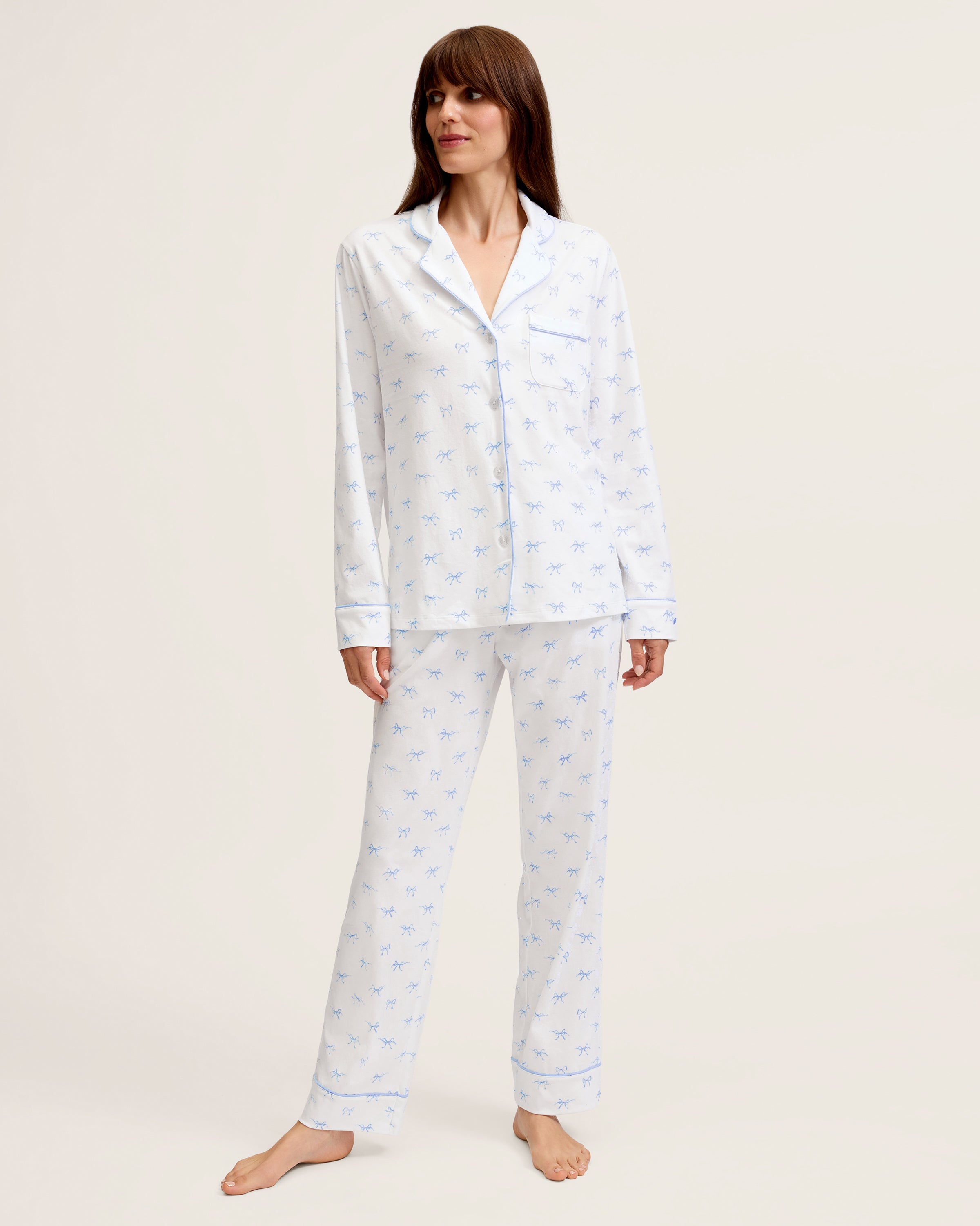 The Women's Pima Pajama Set in Something Blue Bows is a white set with blue piping, including a collared button-up top with chest pocket and matching pants. Both feature a repeating blue print of small bows.