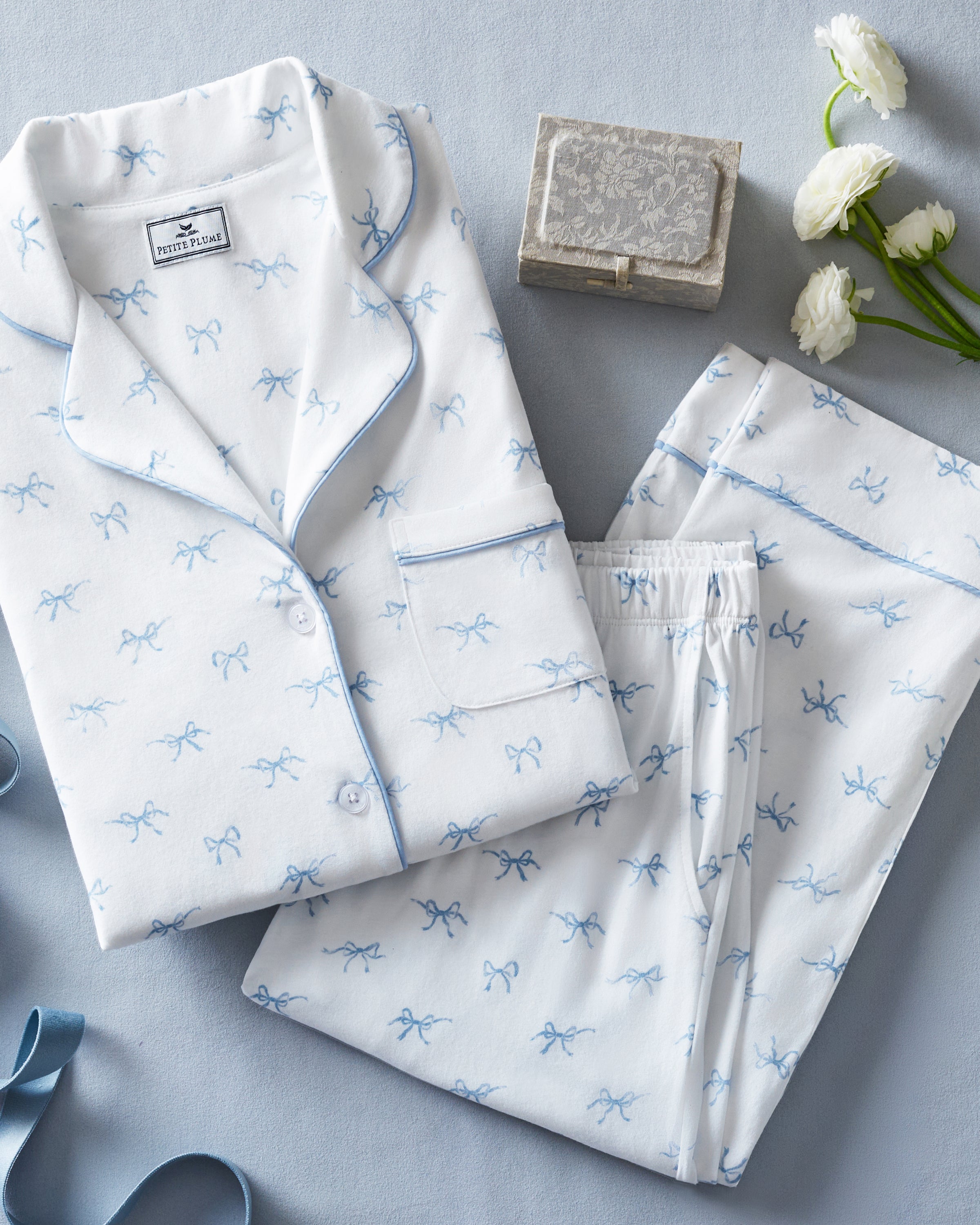 The Women's Pima Pajama Set in Something Blue Bows is a white set with blue piping, including a collared button-up top with chest pocket and matching pants. Both feature a repeating blue print of small bows.