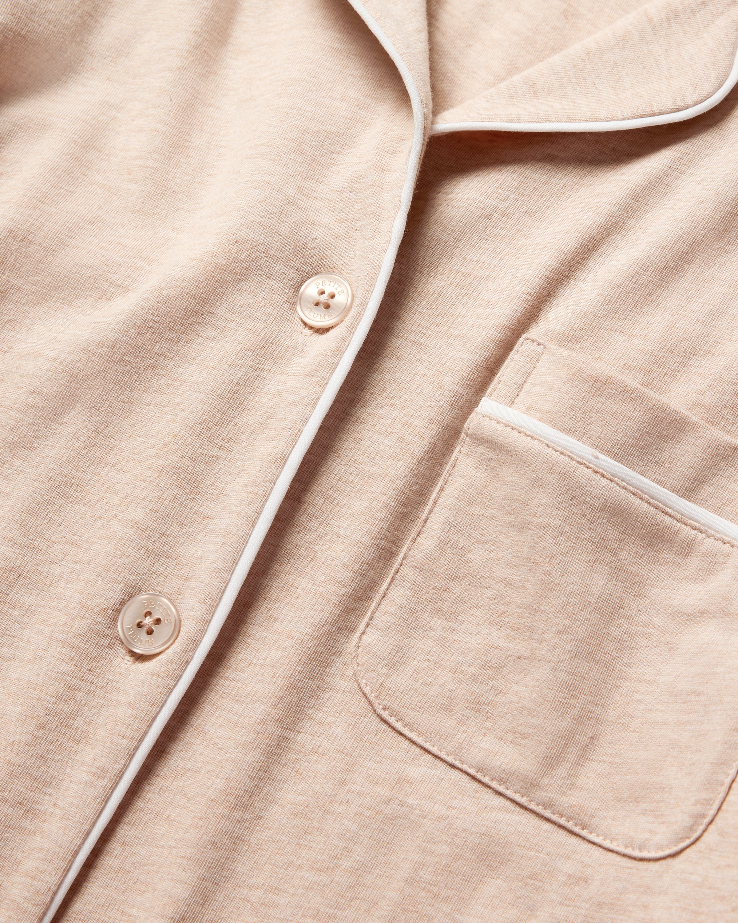 The Womens Pima Pajama Set in Camel by Petite Plume is crafted from soft, textured Pima cotton. It features a close-up of a beige fabric pajama shirt with white piping, a pocket on the right side, and two buttons down the center, showcasing luxury sleepwear at its finest.