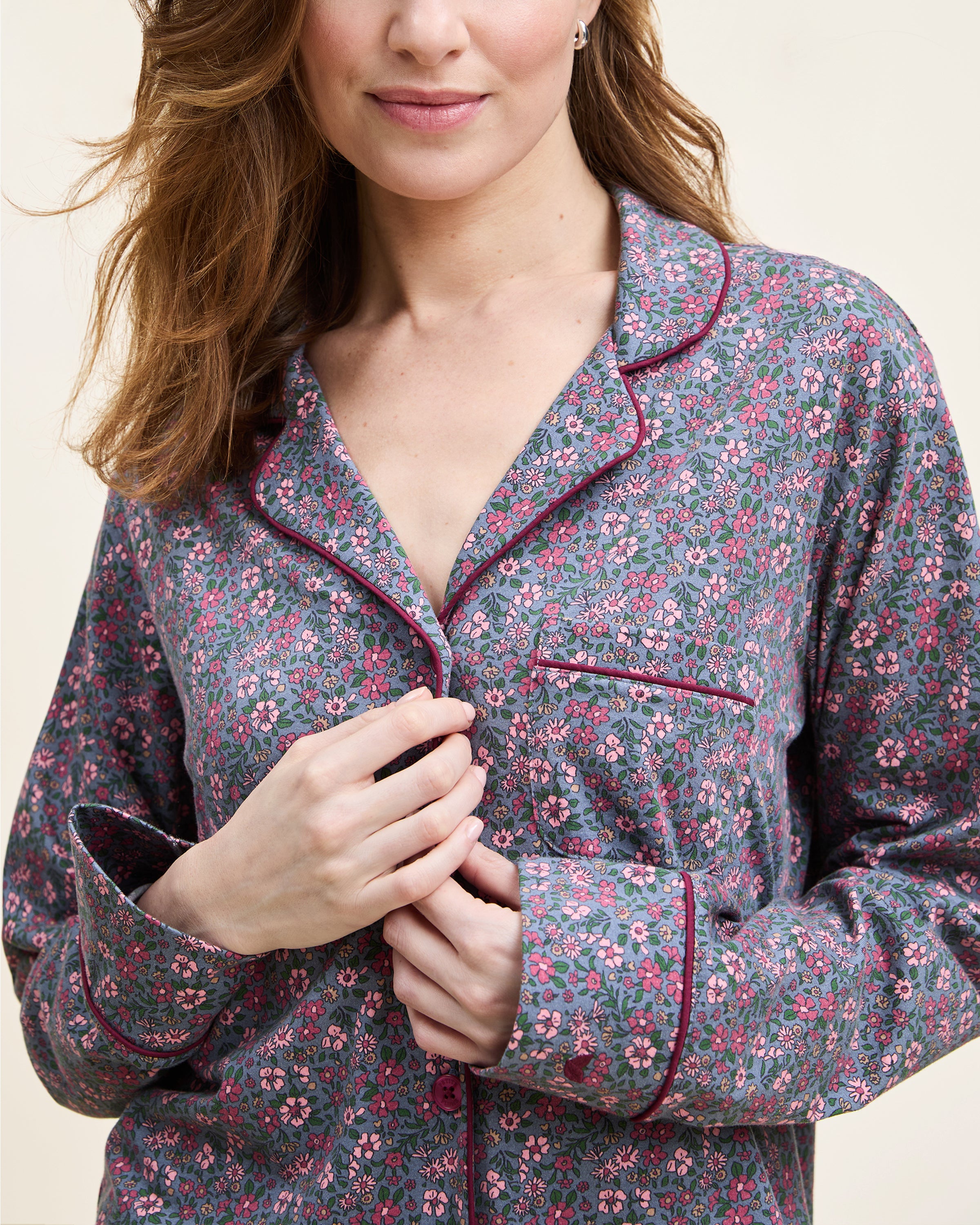 The Women's Pima Pajama Set in Fleurs de Saison features long sleeves and a small floral print in pink, white, and blue on gray. The set includes a button-up top with a chest pocket and matching pants.