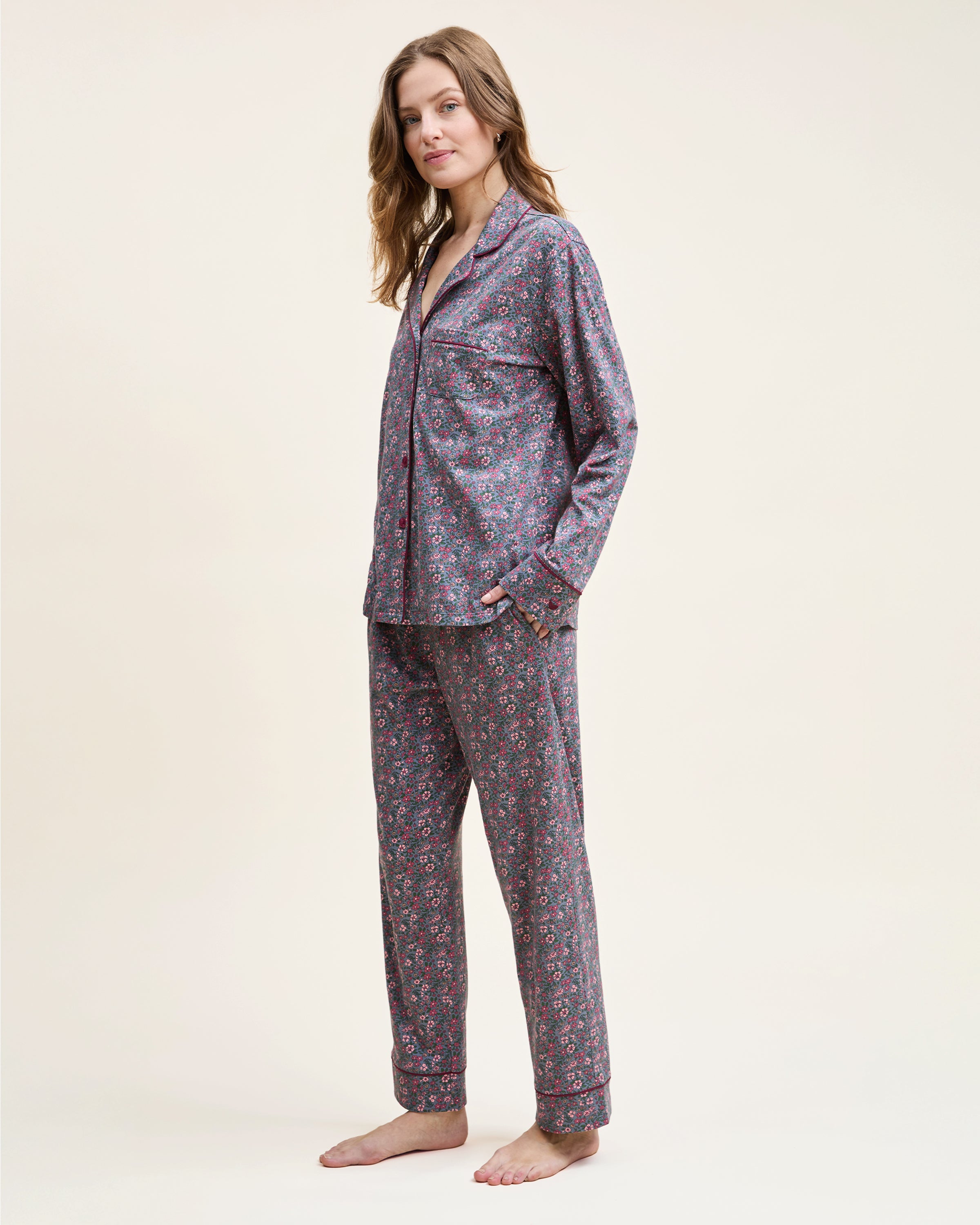The Women's Pima Pajama Set in Fleurs de Saison features long sleeves and a small floral print in pink, white, and blue on gray. The set includes a button-up top with a chest pocket and matching pants.