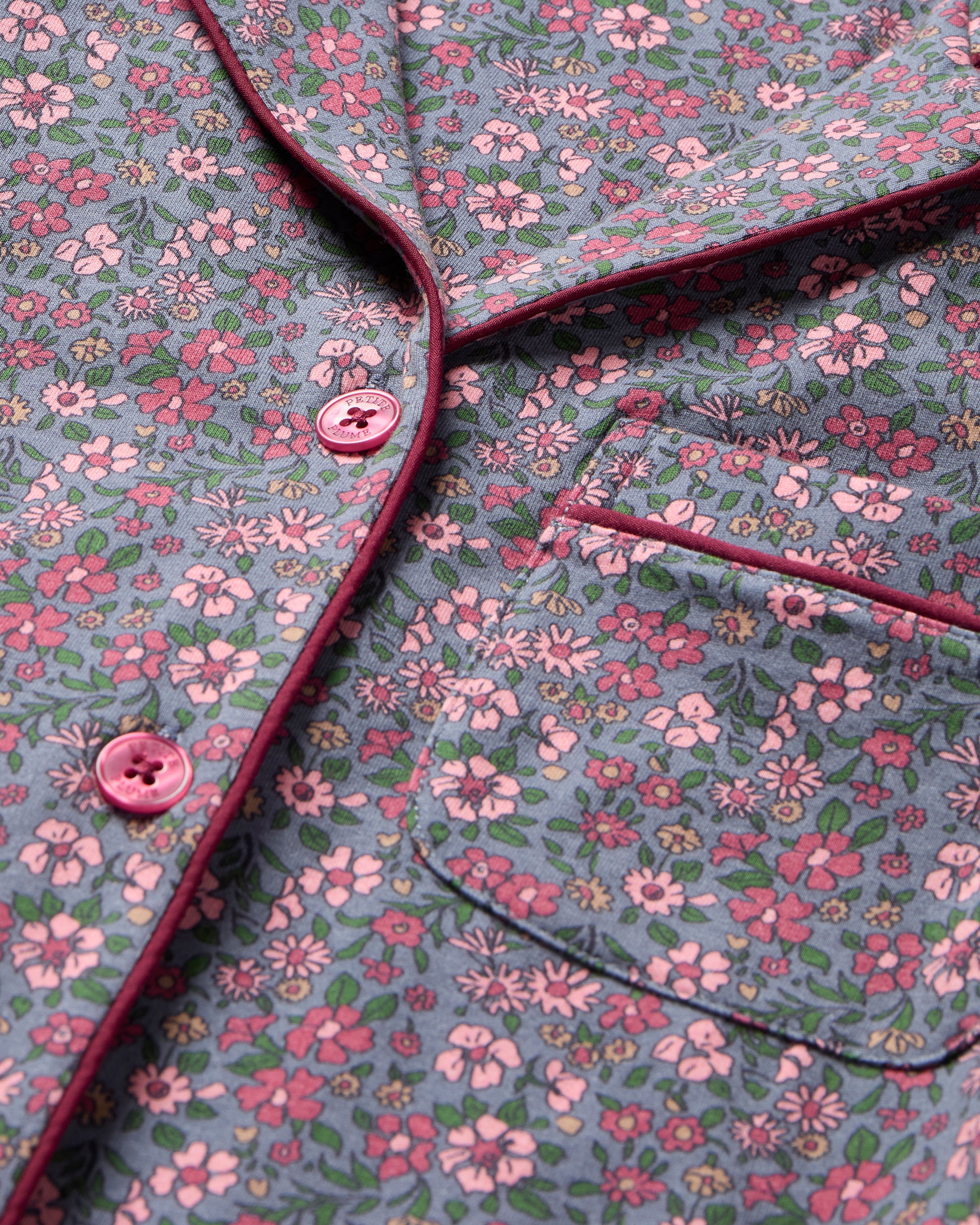 The Women's Pima Pajama Set in Fleurs de Saison features long sleeves and a small floral print in pink, white, and blue on gray. The set includes a button-up top with a chest pocket and matching pants.