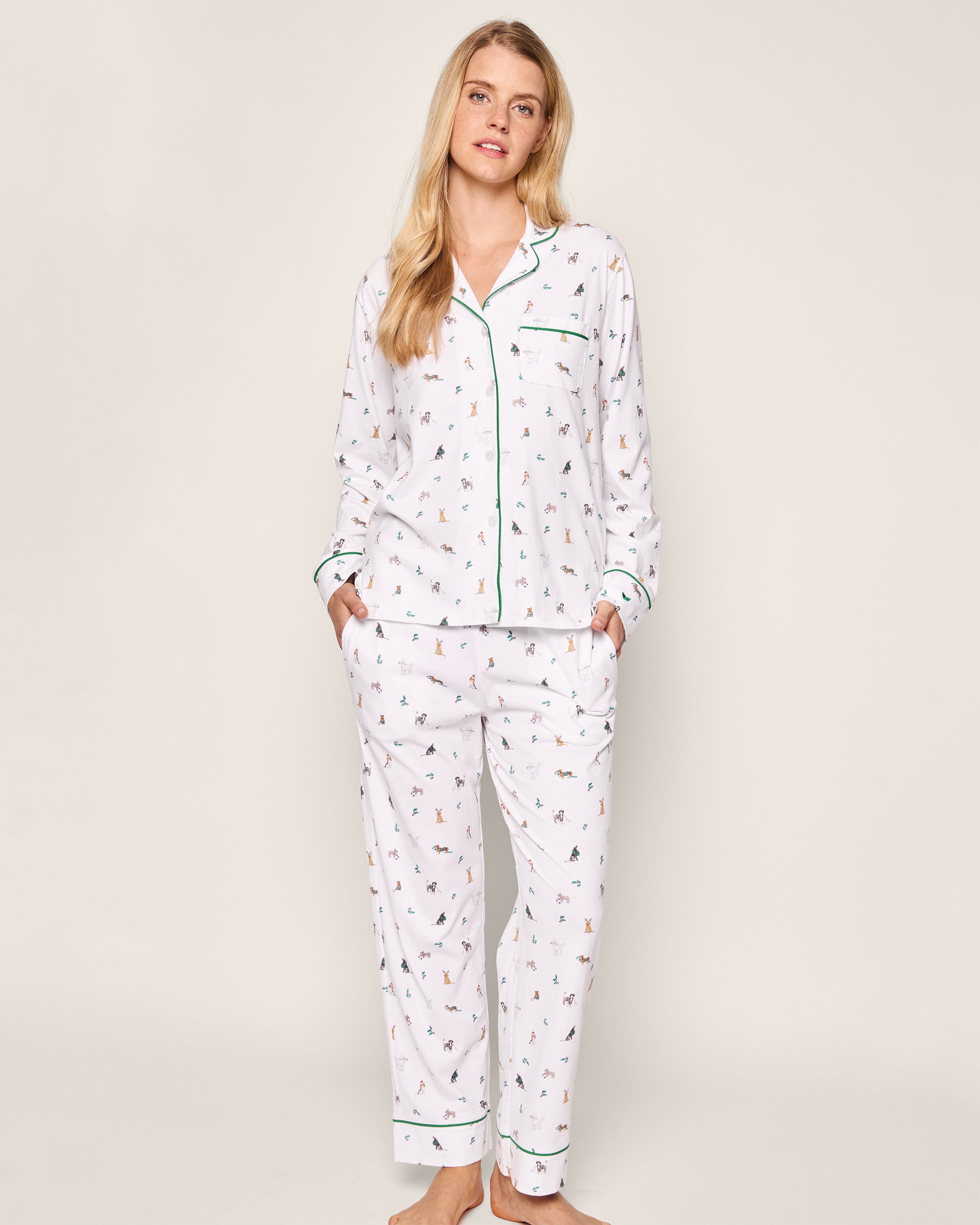 Dressed in Petite Plumes Womens Pima Pajama Set in Jingle Paws, an individual with long hair stands against a neutral backdrop. The luxurious pajamas, accented with green piping, feature an animal print and embody holiday sleepwear elegance as they place hands in pockets.