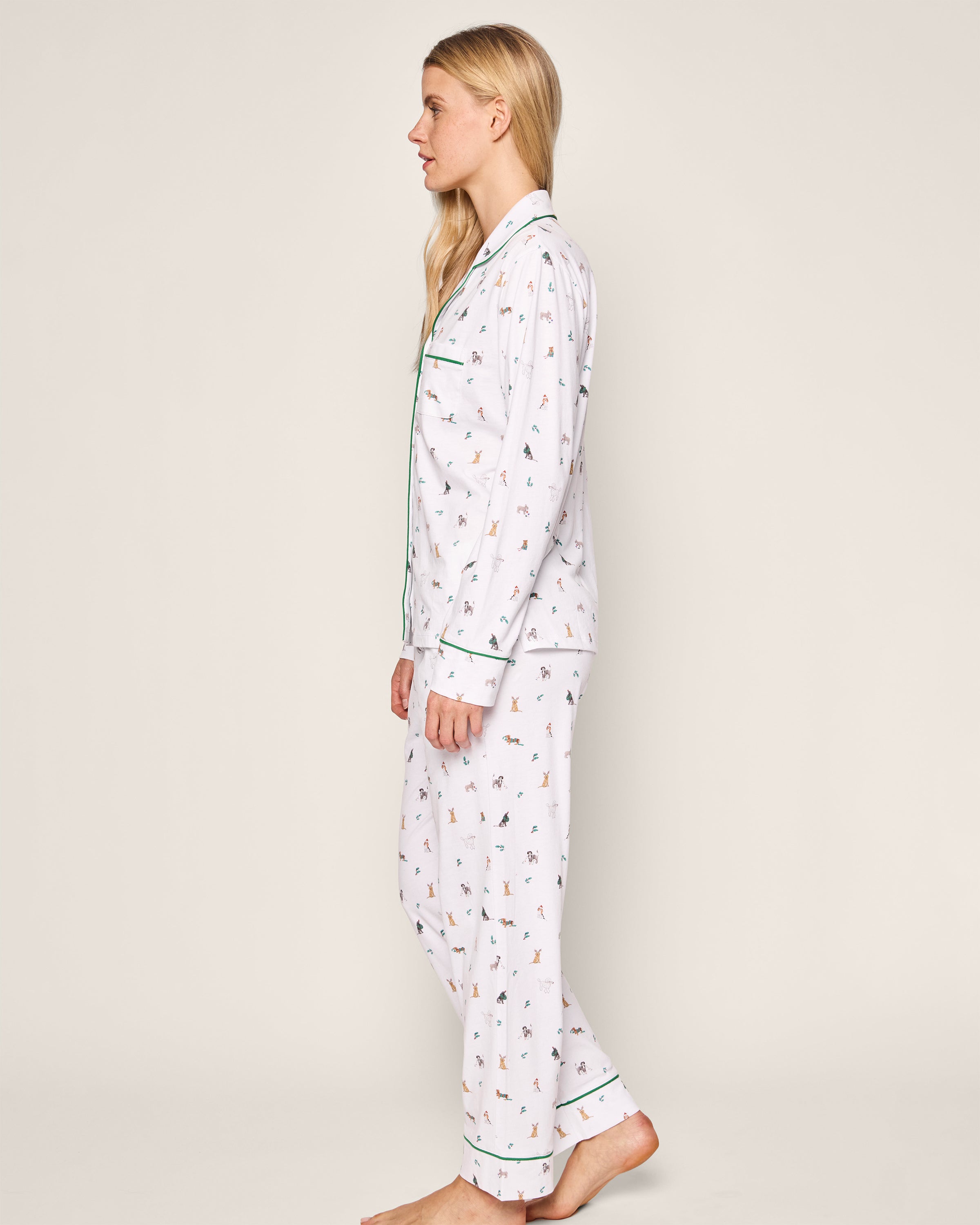 A person with long blonde hair stands barefoot in profile, wearing the Womens Pima Pajama Set in Jingle Paws by Petite Plume. The set features a playful print, green trim, and a button-up top with small colorful figures, all against a plain white background.