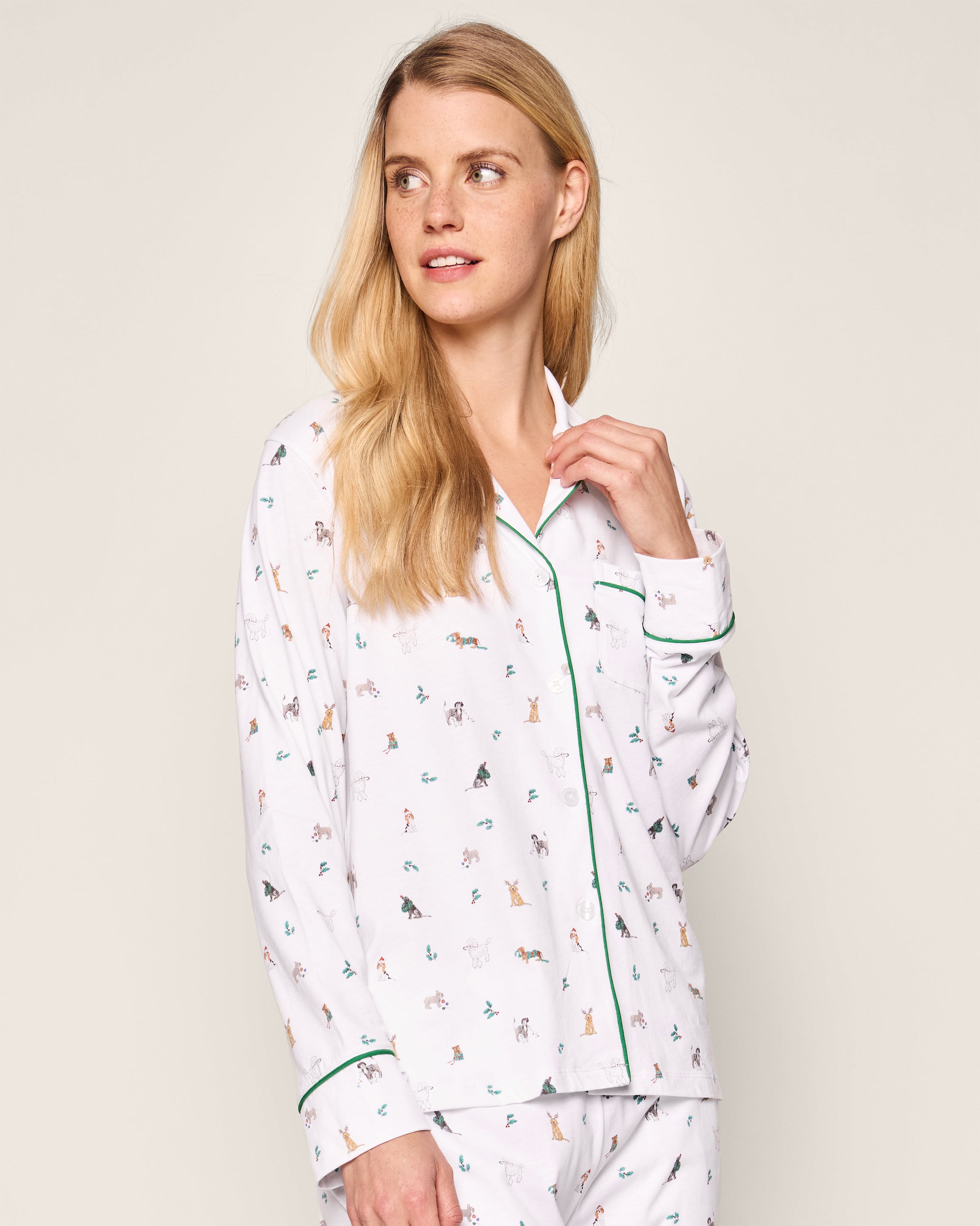 A blonde person wears Petite Plumes Womens Pima Pajama Set in Jingle Paws, crafted from luxurious Pima cotton with playful animal prints and green piping. Theyre slightly turned to the side, showing a neutral expression against a plain background, exuding holiday sleepwear elegance.