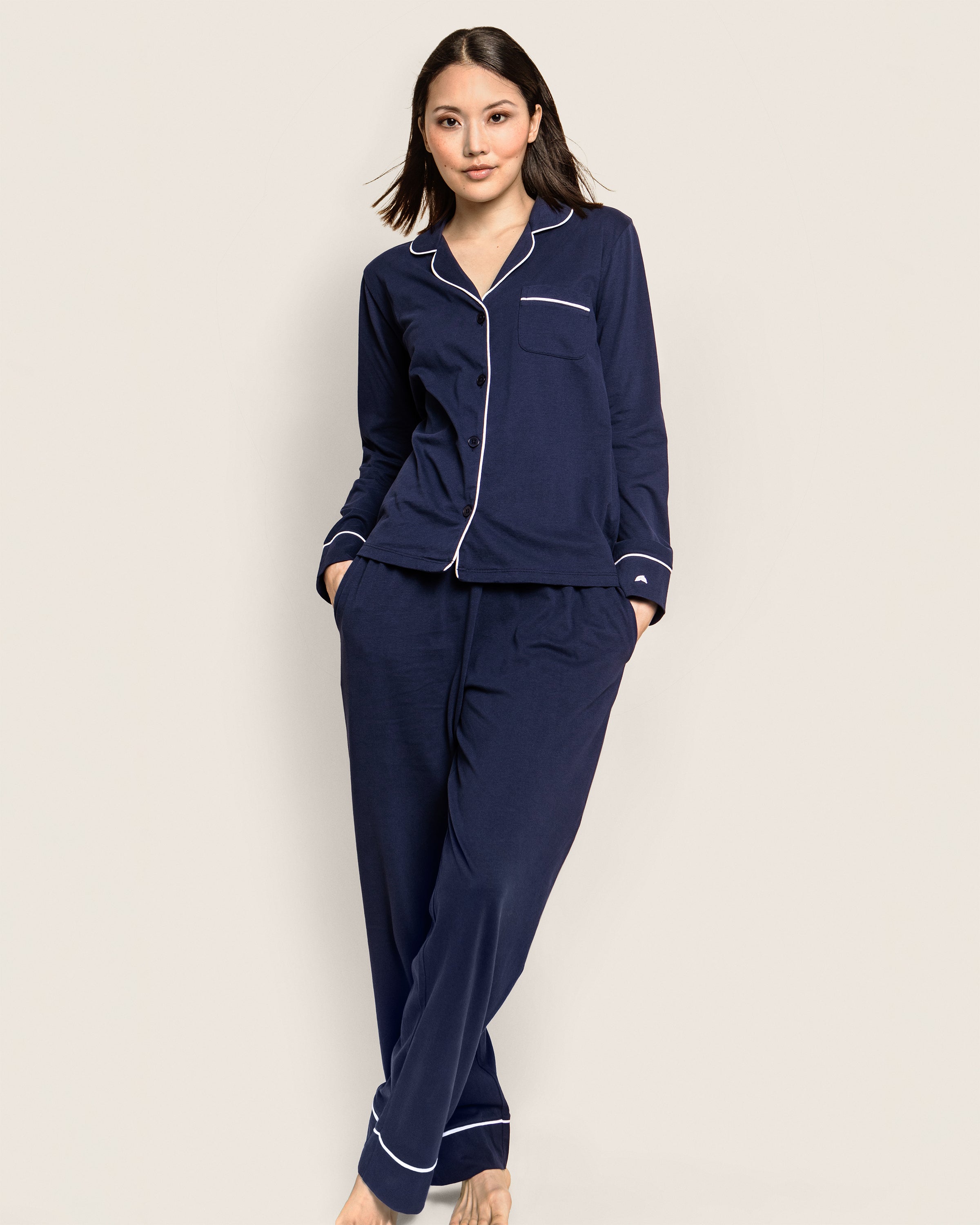 A person models the Petite Plume Womens Pima Pajama Set in Navy, featuring white piping and crafted from luxurious Pima cotton. The button-up top and pants offer ultimate comfort, available in plus sizes. They stand barefoot against a plain, light background, exuding relaxed elegance.